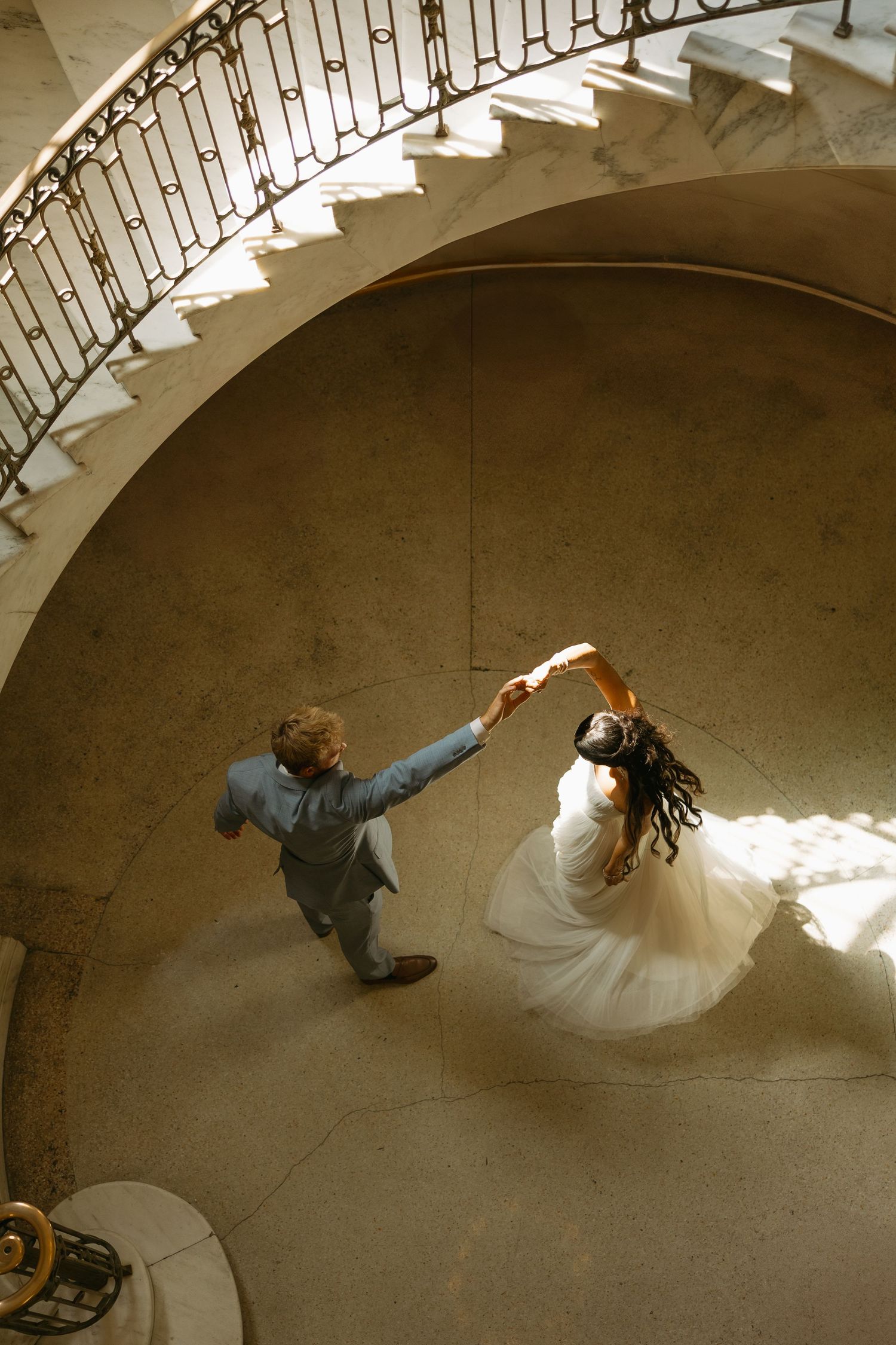 A couple in wedding attire dances beneath a curved staircase in a dramatic overhead view sequence.