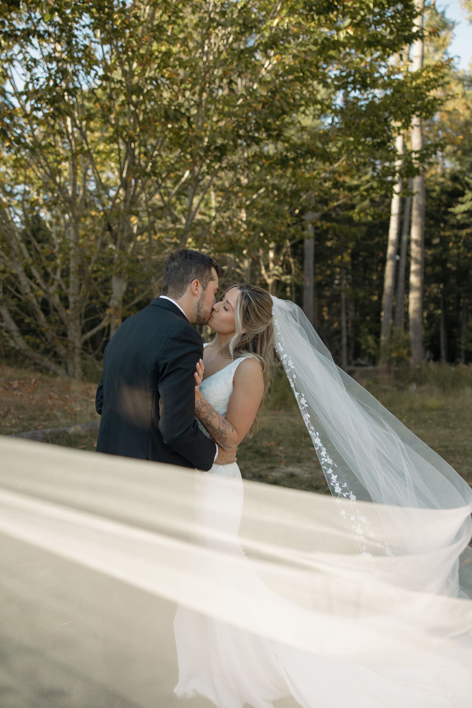 A romantic wedding couple shares a tender kiss outdoors with a flowing white veil in the sunlit forest.