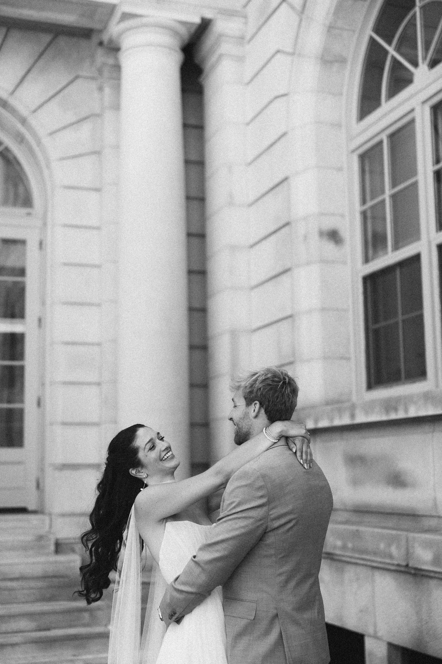 A couple shares a romantic moment against a classical building facade with white columns and arched windows.