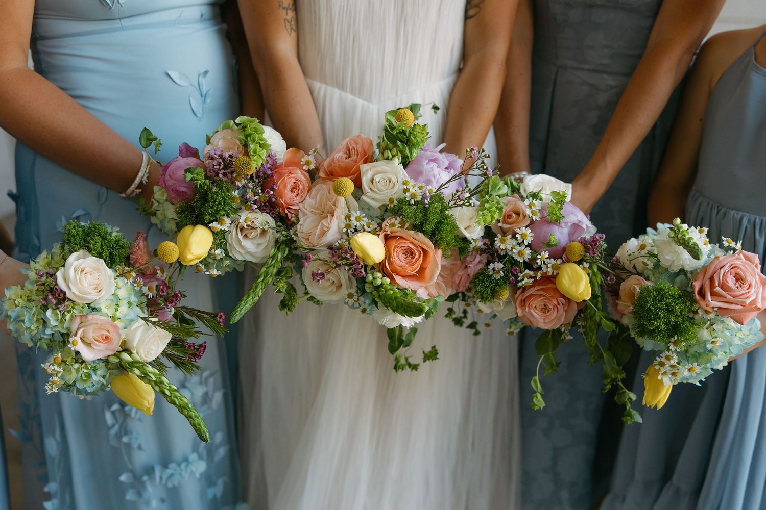 Bridesmaids in blue dresses hold colorful bouquets of pink, yellow and white flowers against a white background.