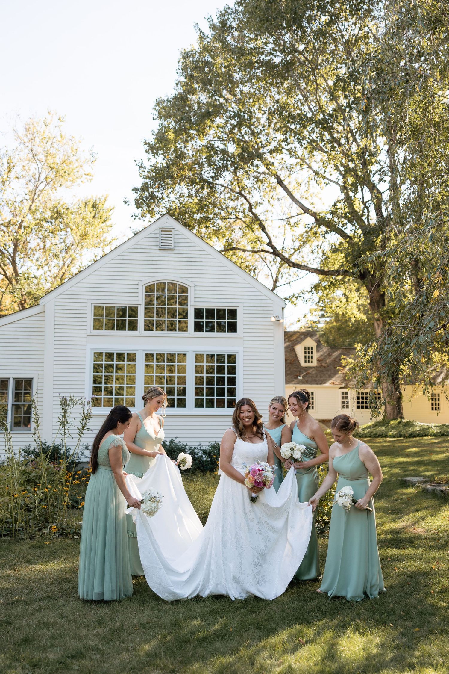 A wedding party in mint green dresses poses playfully in front of a white barn building on a sunny day.