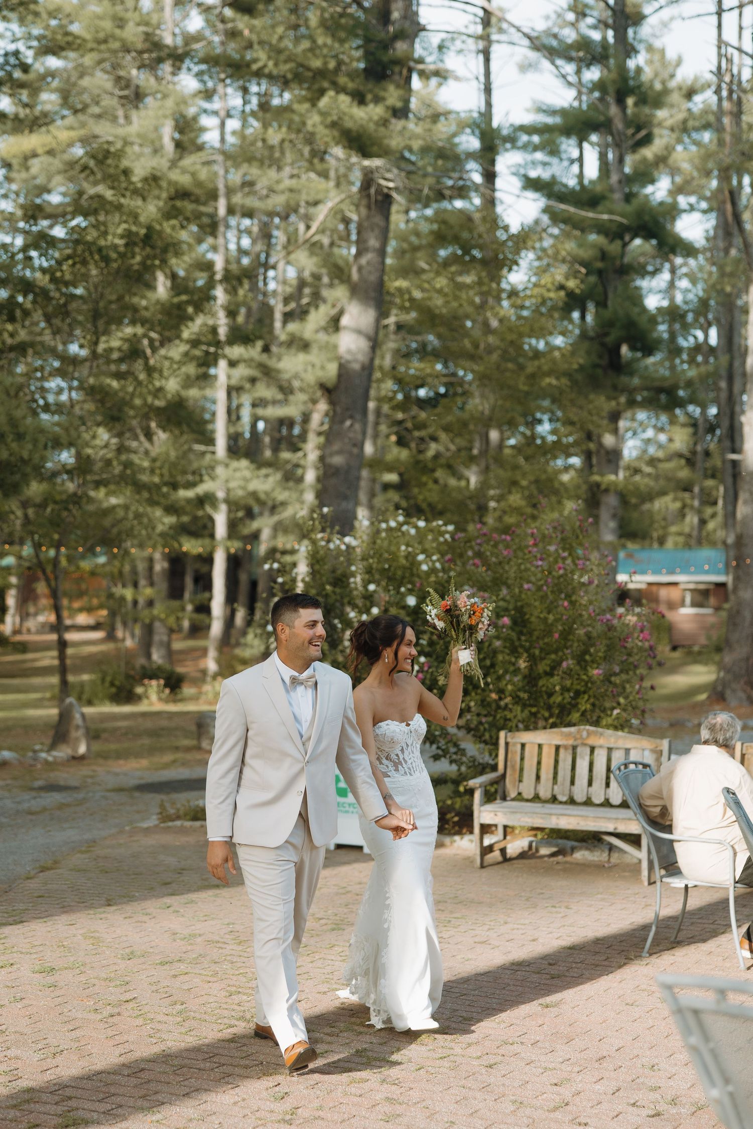 A couple celebrates their wedding day walking along a brick path surrounded by tall pine trees at an outdoor venue.