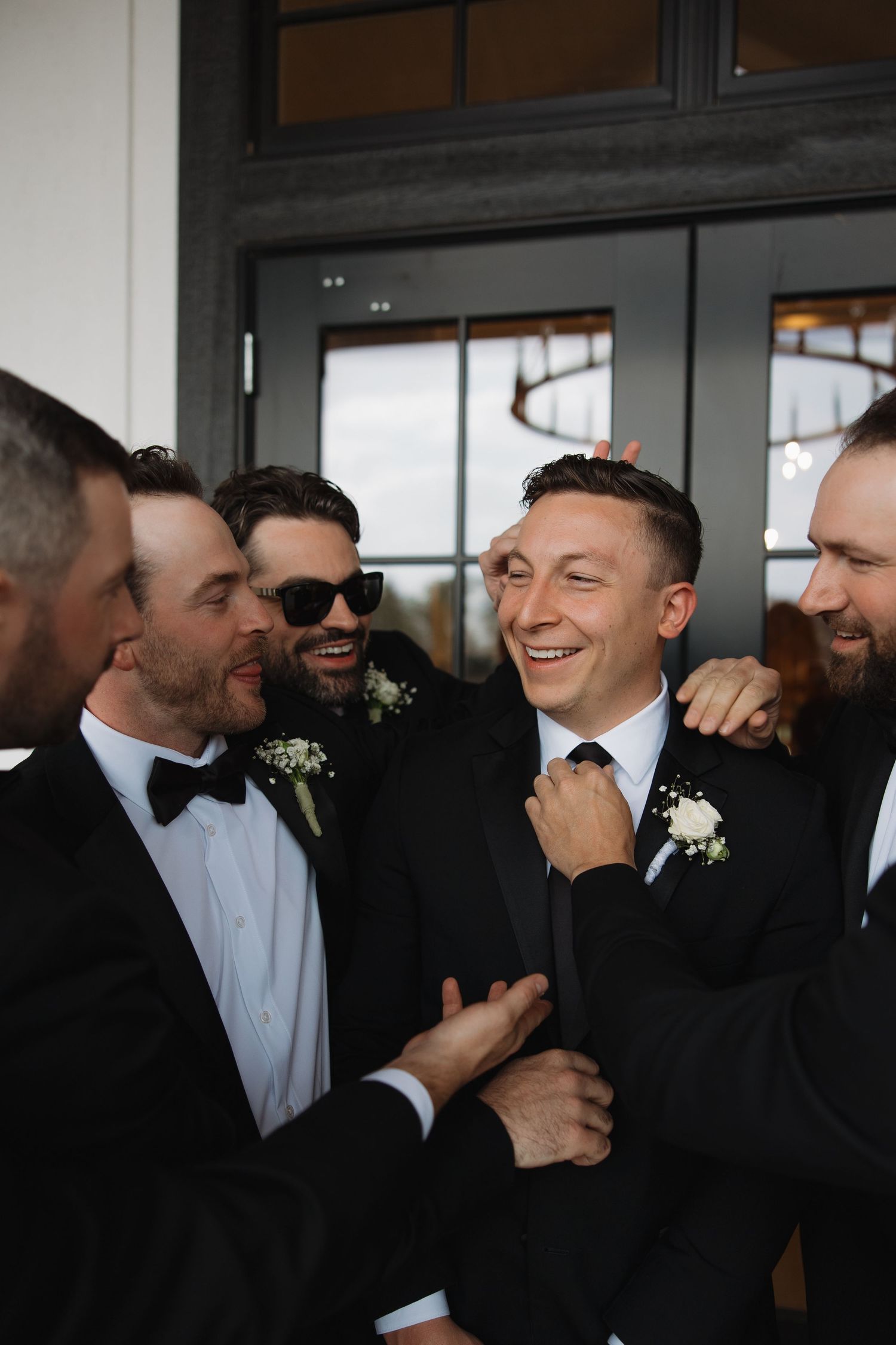 A candid sequence of groomsmen sharing laughter and embraces while wearing black tuxedos and white shirts at a wedding.