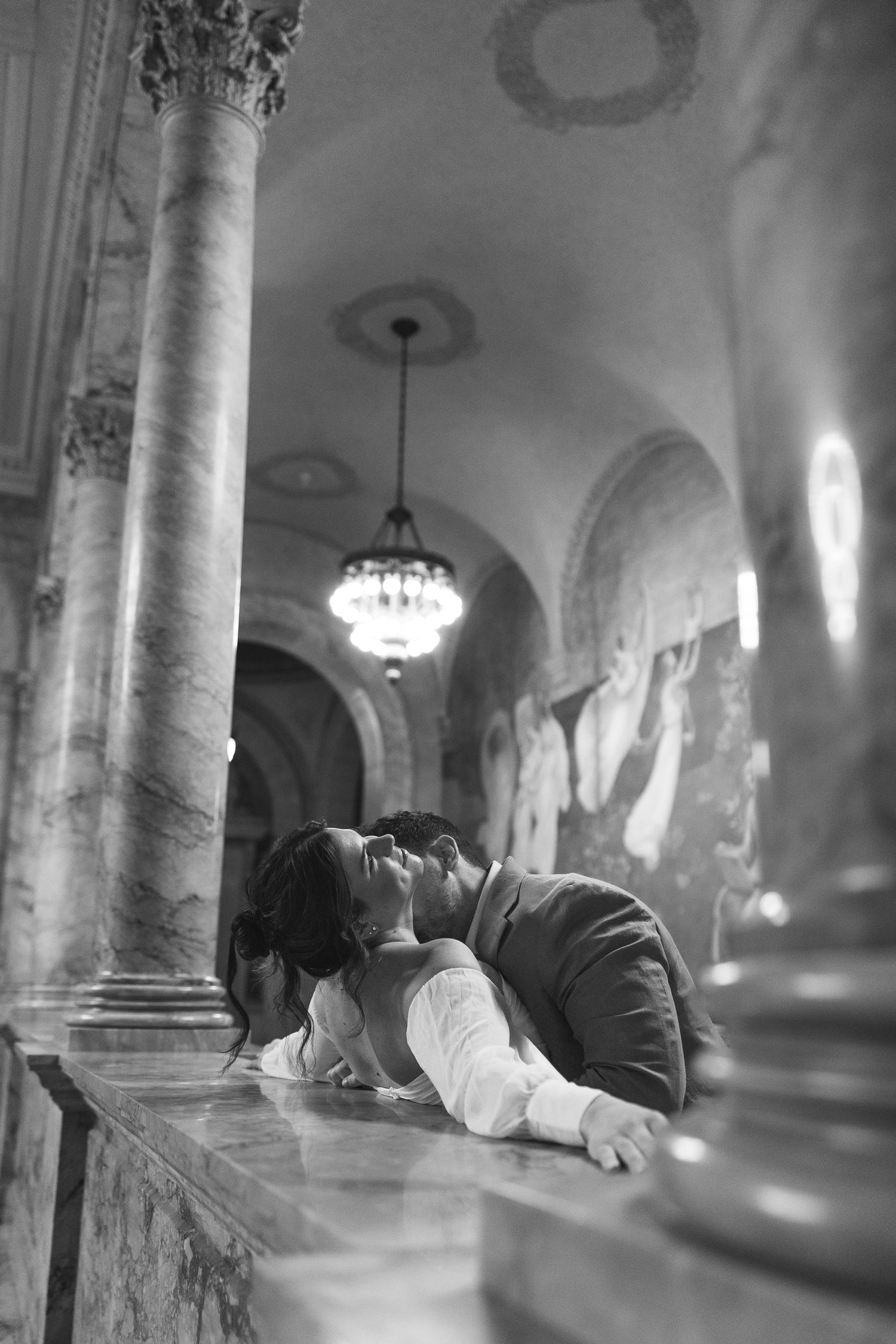 A person lies sleeping on a stone ledge inside a grand historic building with ornate columns and chandeliers.