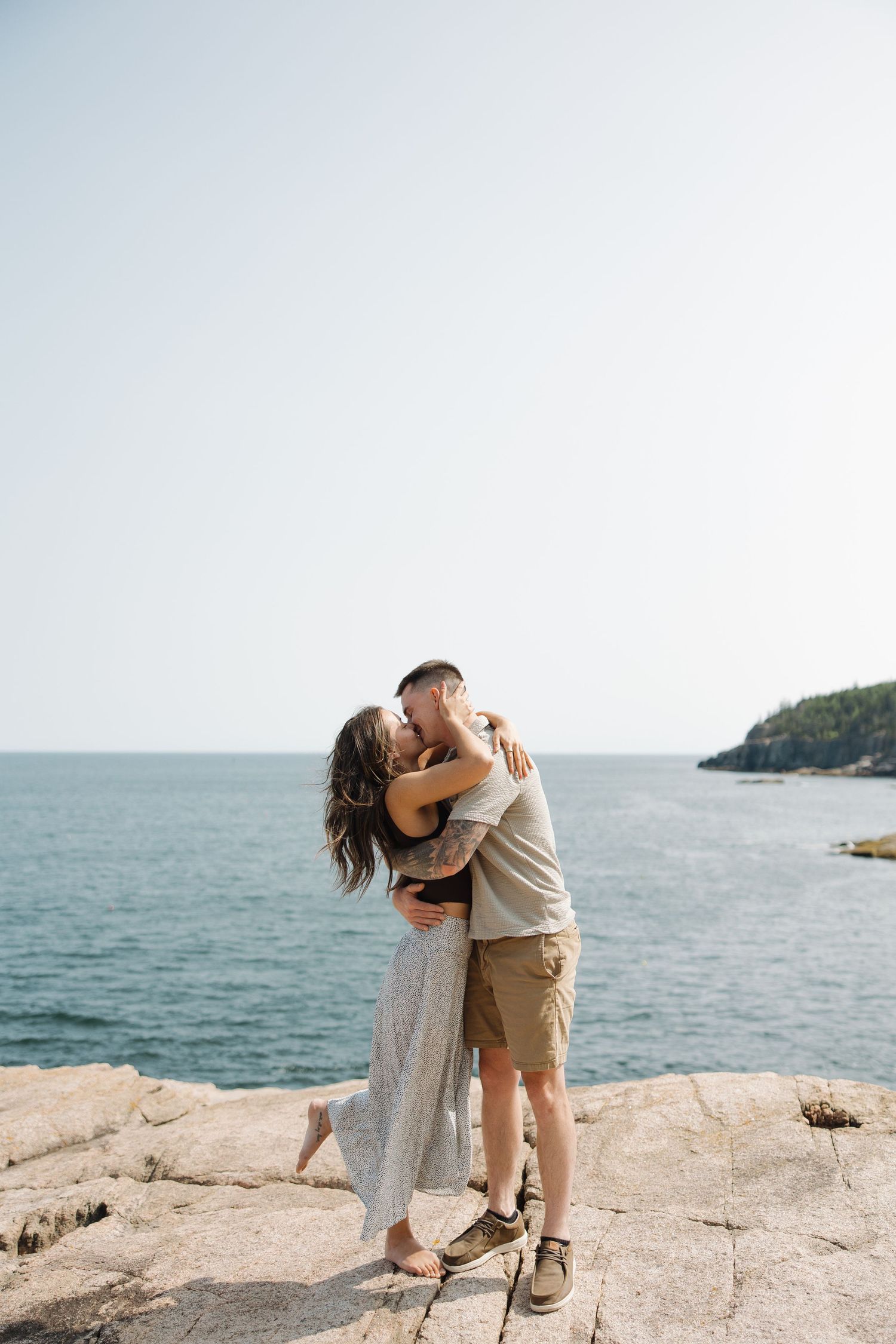 A romantic couple embraces on rocky coastal cliffs overlooking a serene ocean view on a bright sunny day.