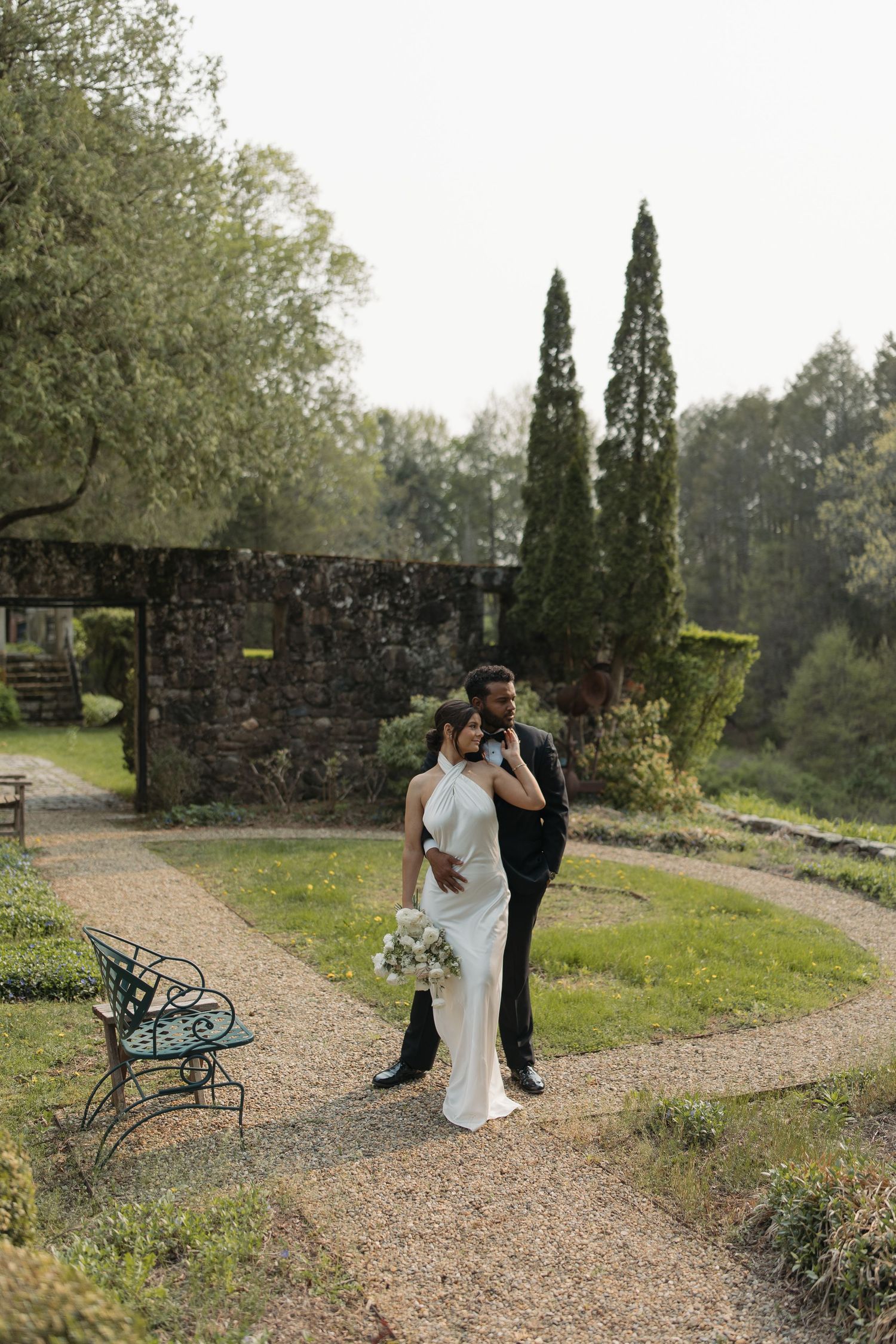 A couple walks along a scenic gravel path surrounded by cypress trees and stone walls in a romantic garden setting.