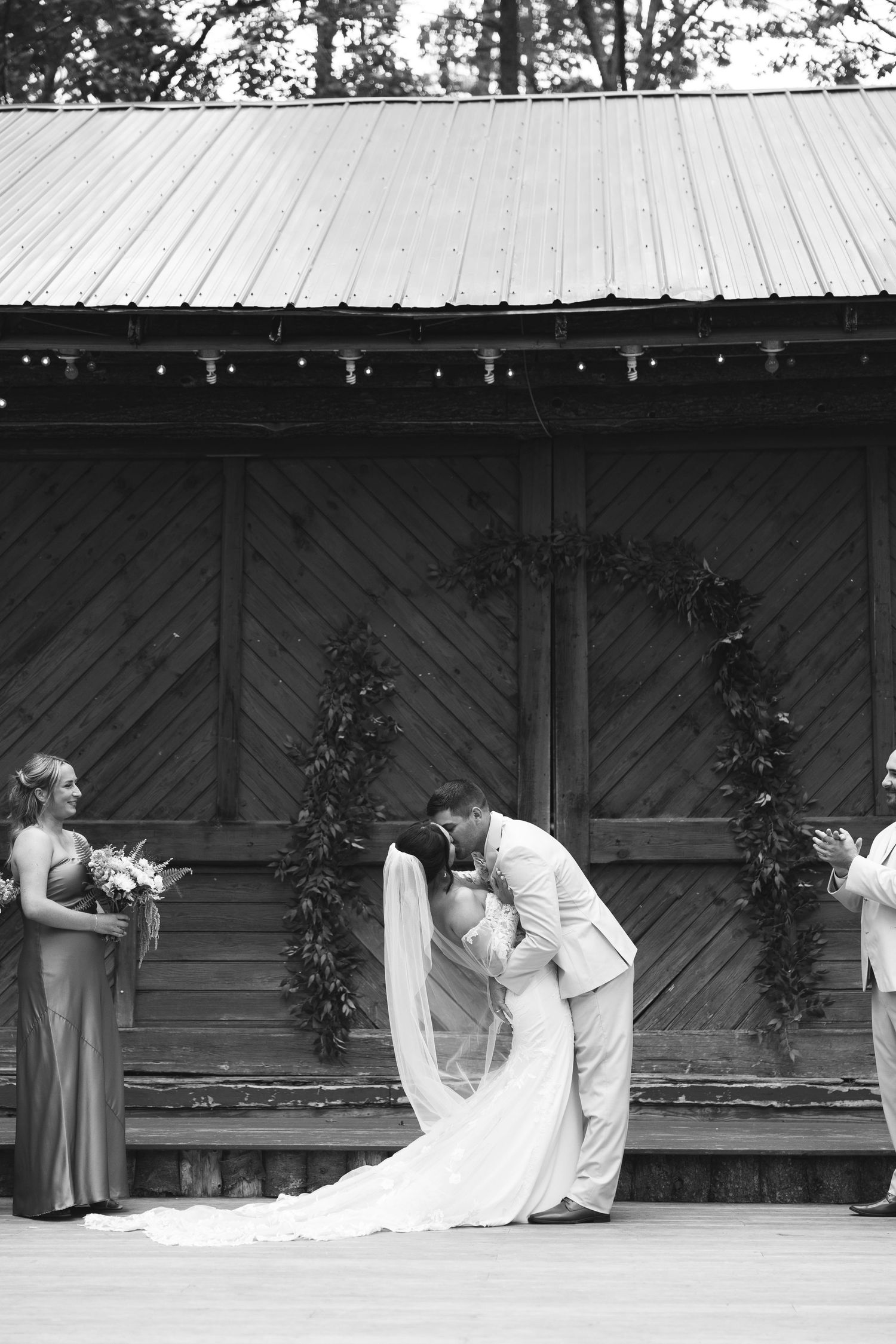 A romantic wedding moment captured in black and white at a rustic wooden pavilion with string lights and greenery.