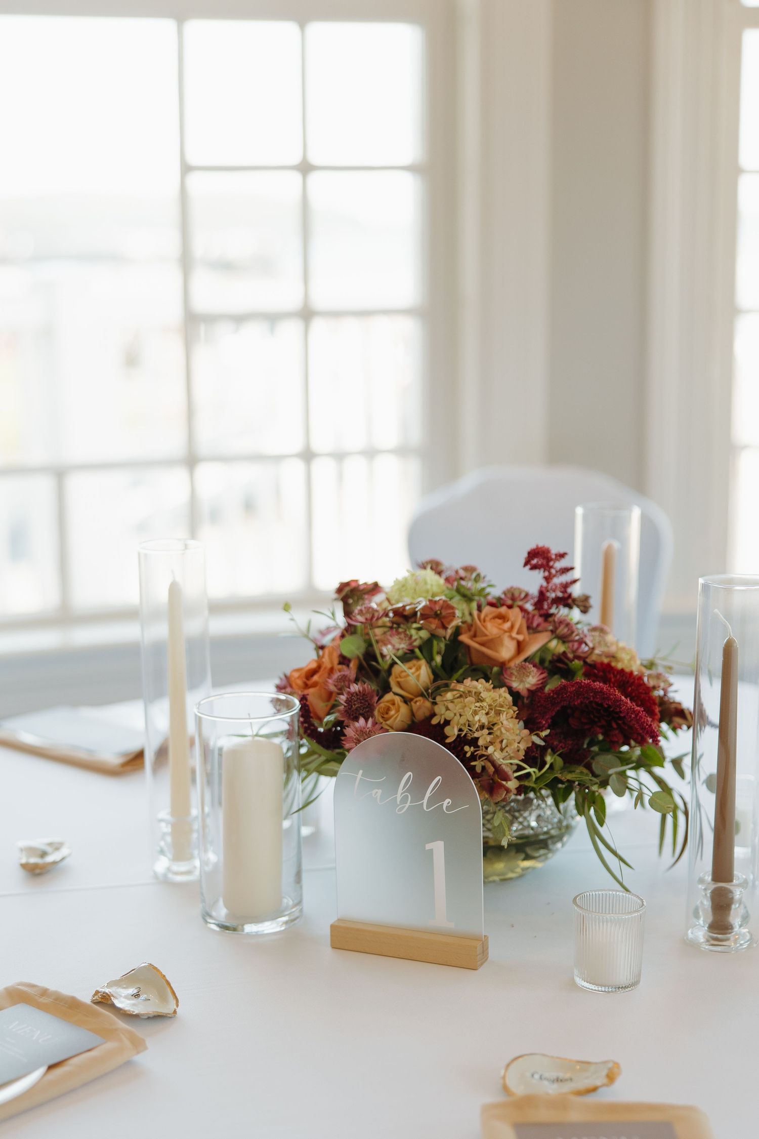 Elegant white table setting with floral centerpieces featuring burgundy and peach blooms alongside glass candle holders.
