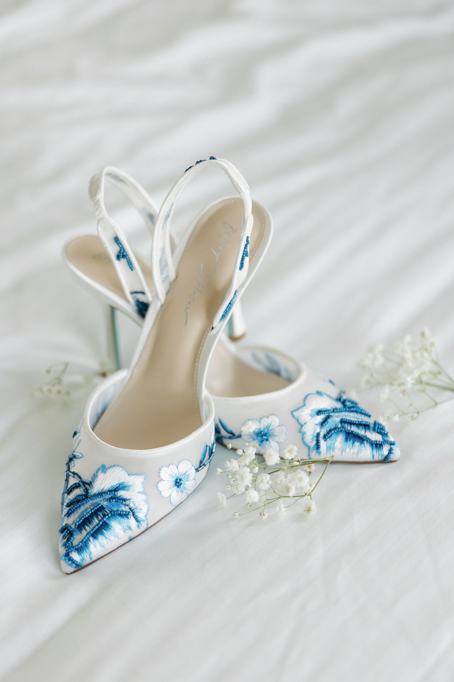 Elegant white bridal shoes with delicate blue floral patterns and small flowers displayed on white fabric.