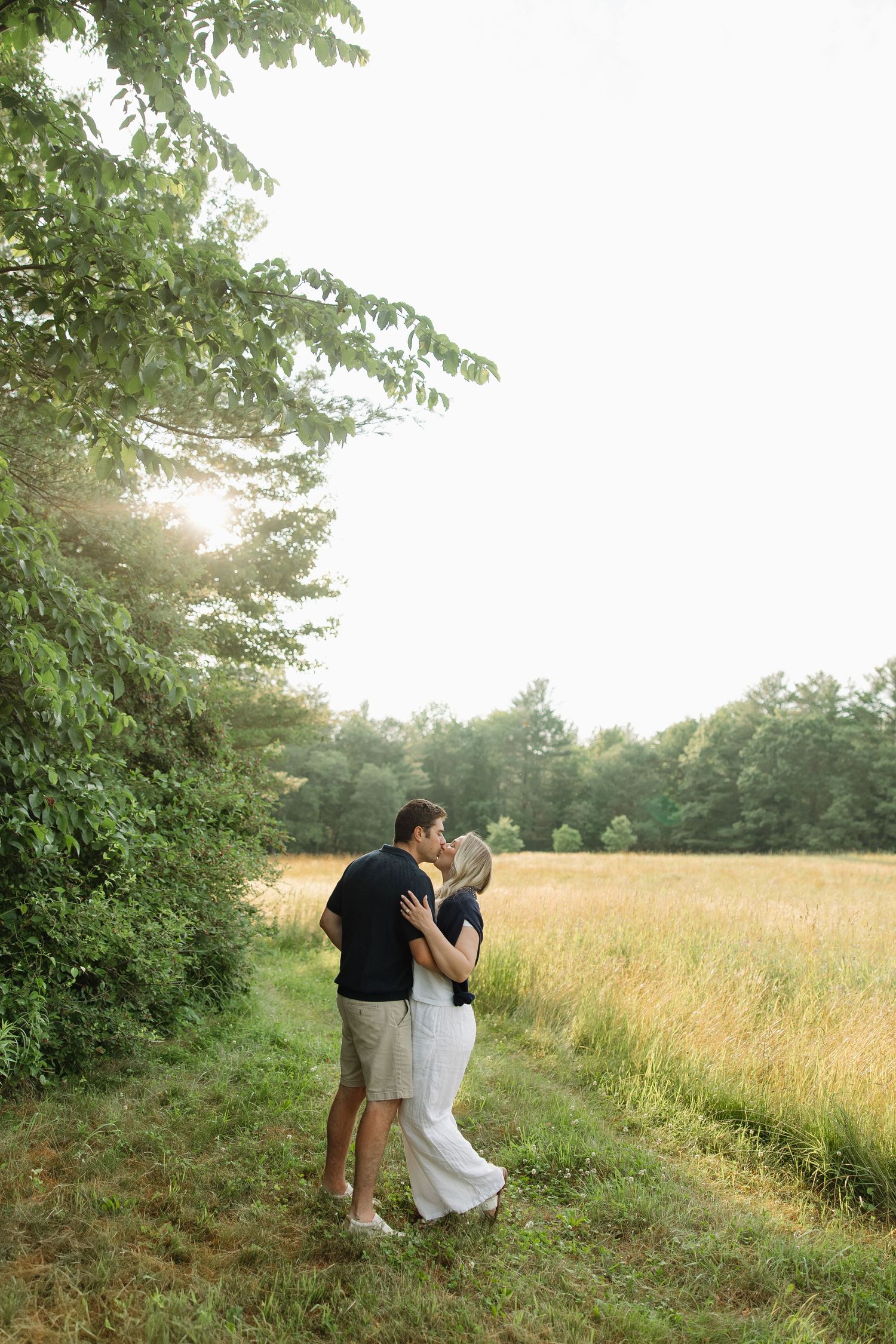 A couple shares a romantic moment in a sunlit meadow with tall grass and trees in the background.