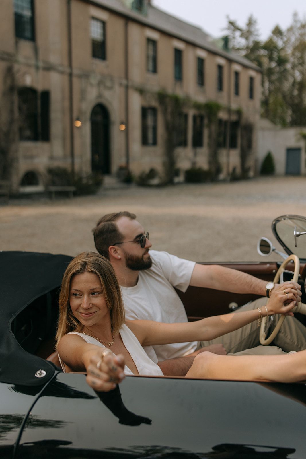 A woman in a white dress poses playfully on a vintage car in an urban setting with stone buildings in the background.