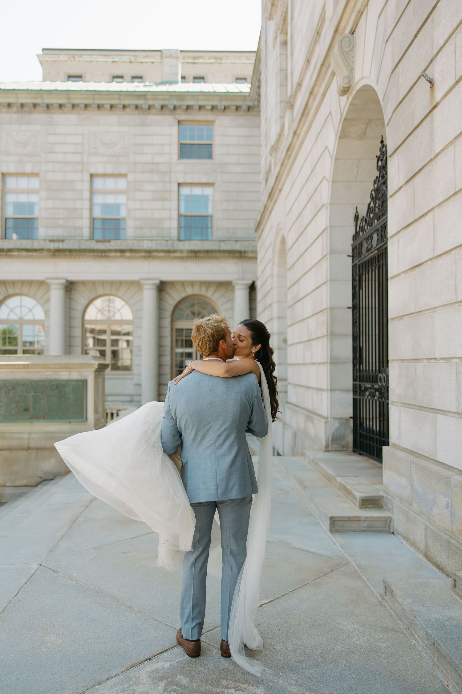 A couple in wedding attire walk along an elegant white stone building exterior with arched windows and columns.