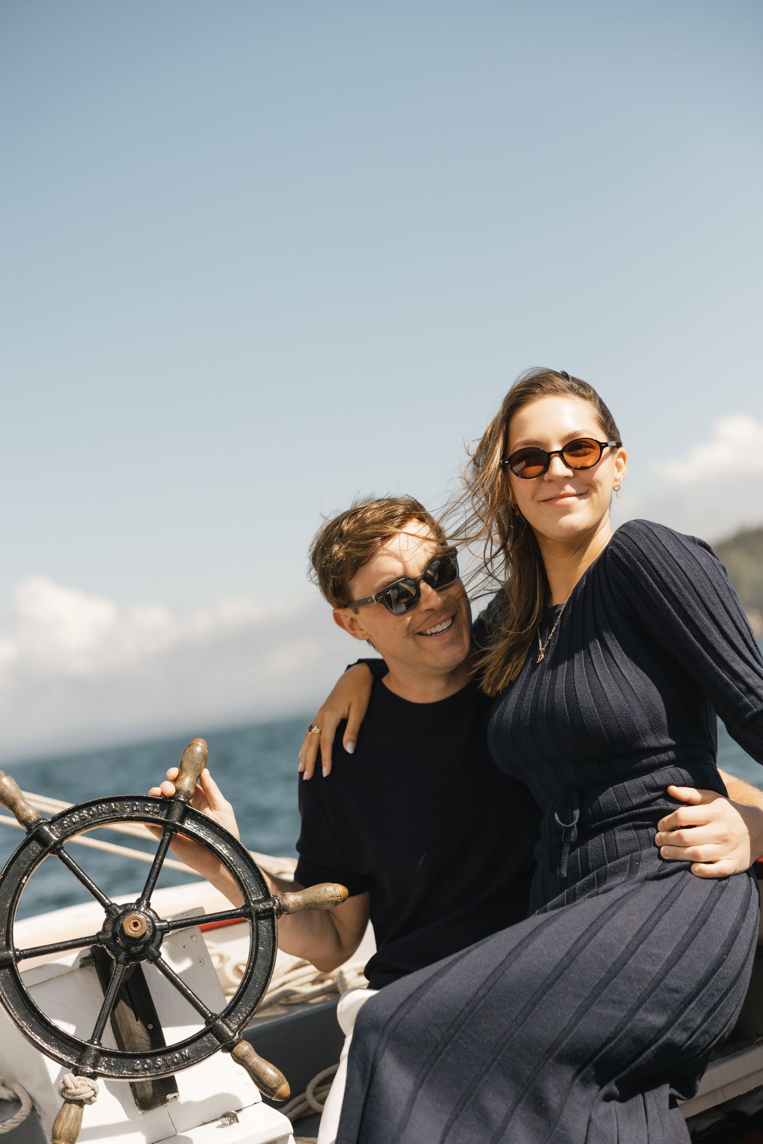 A couple wearing black outfits enjoys a sunny day on a boat, sitting at the helm and steering wheel.