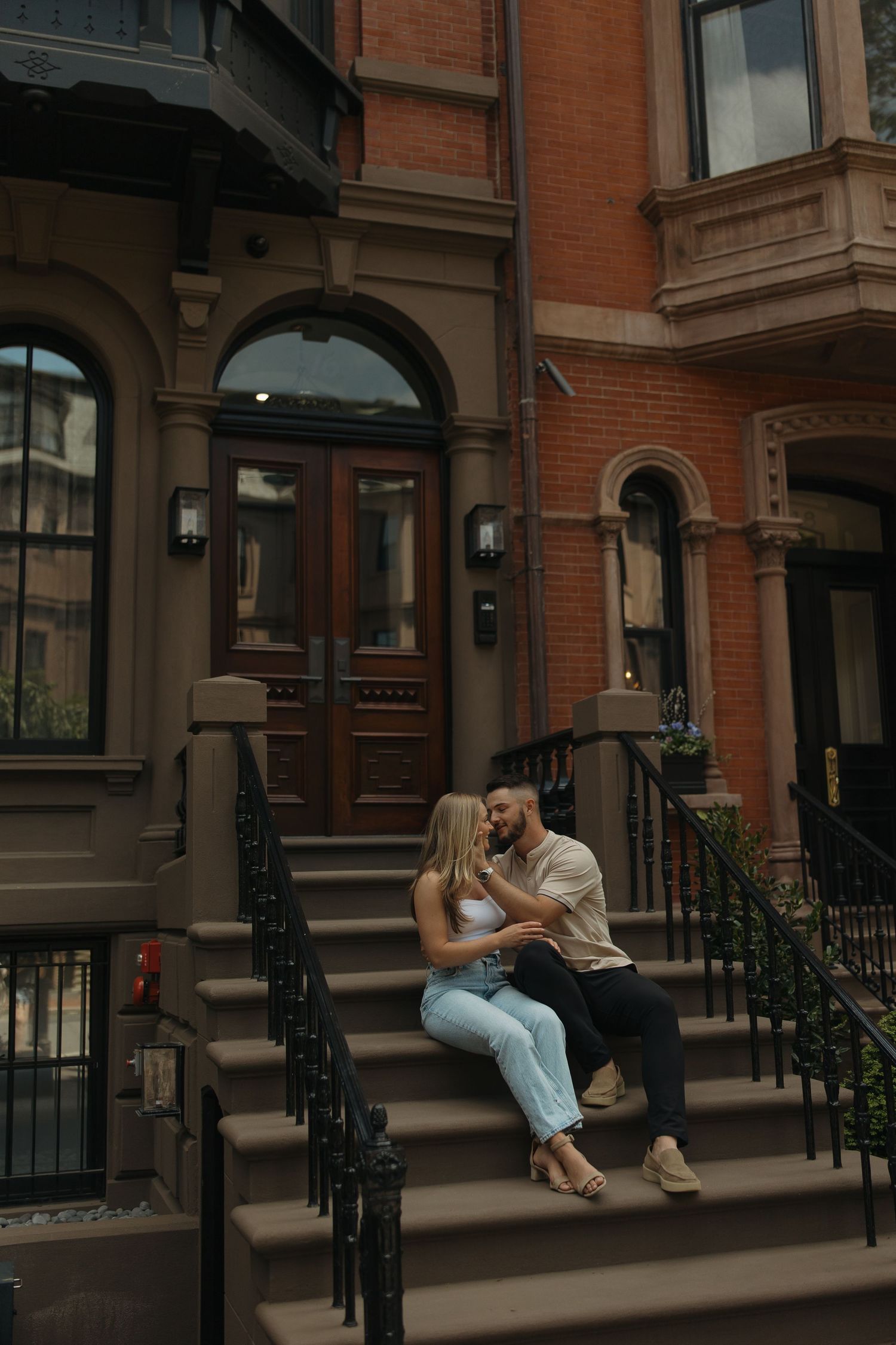A couple sits intimately on brownstone steps in front of a classic brick building with arched windows and iron railings.