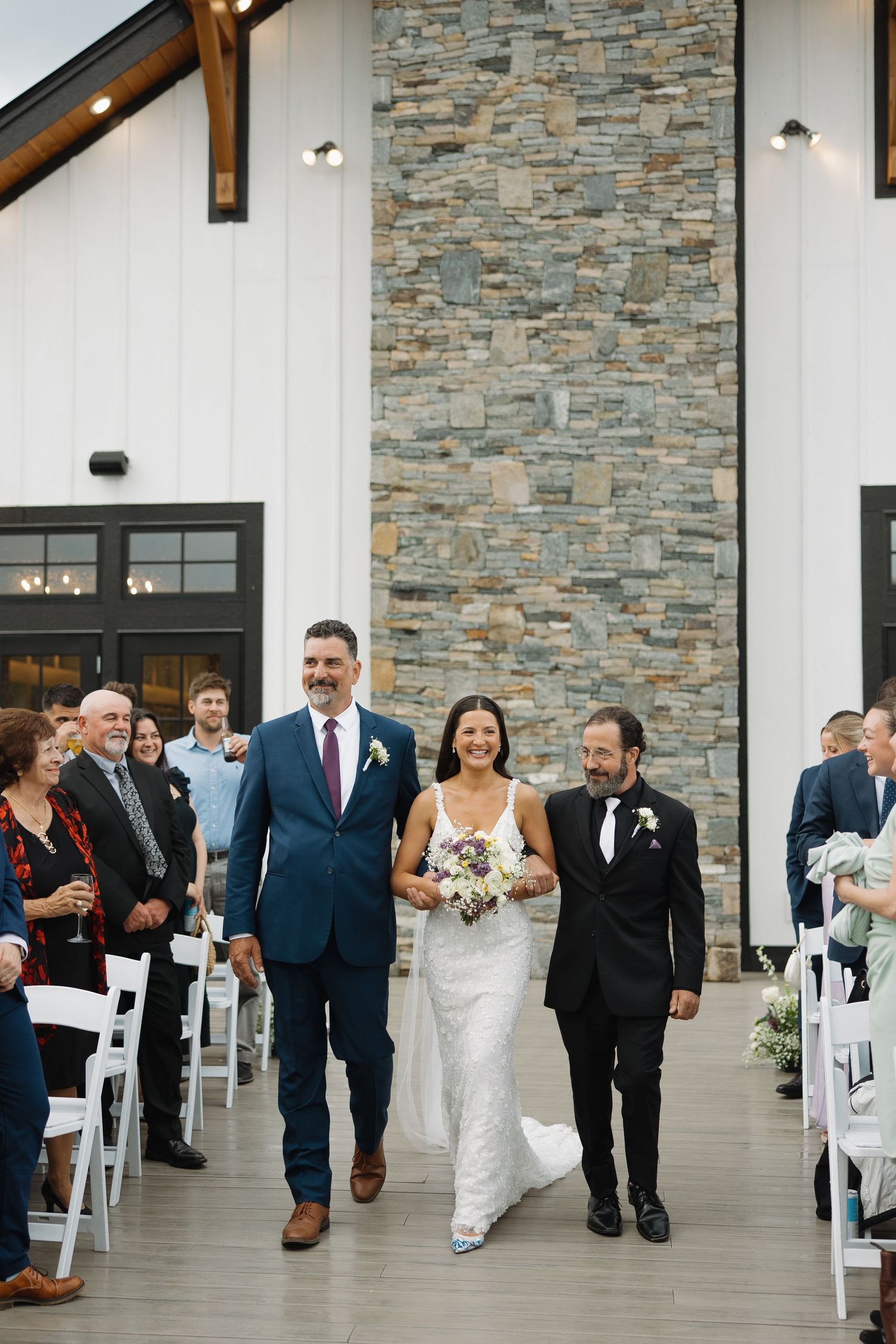 Wedding party walks down outdoor covered walkway with stone walls and white chairs during ceremony recessional.
