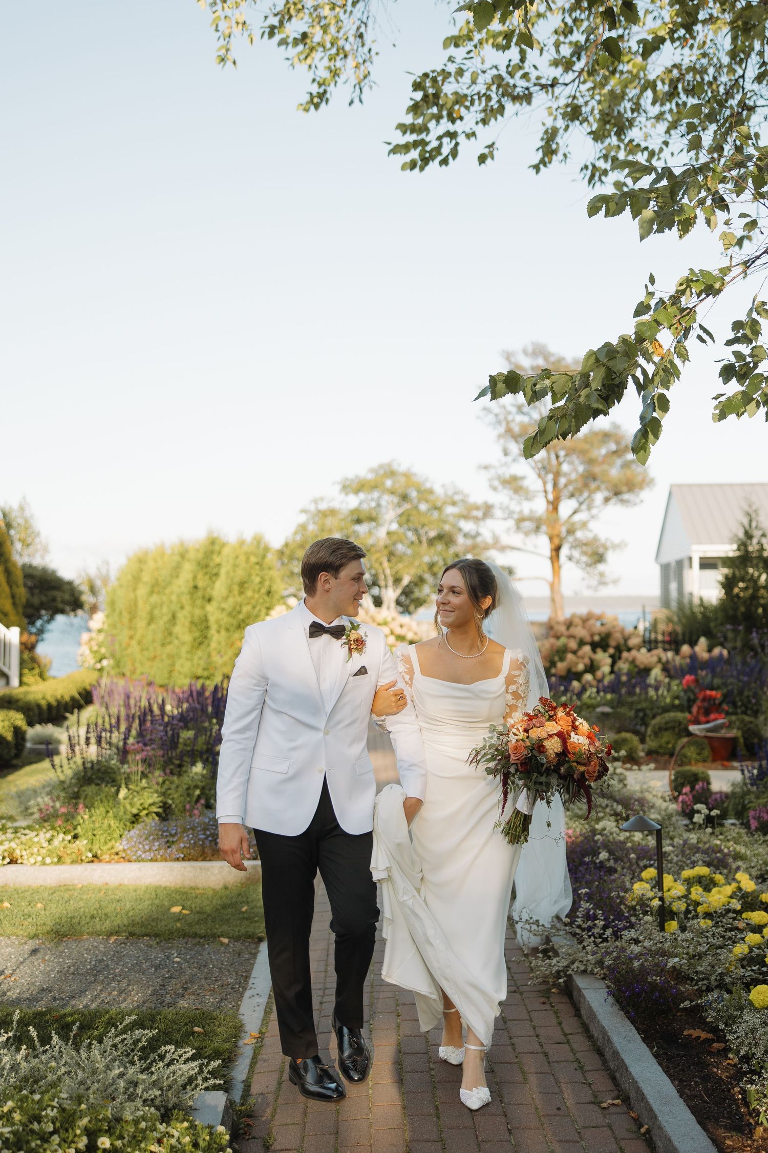 A couple walks together through a lush garden pathway with colorful flowers and greenery on a sunny day.