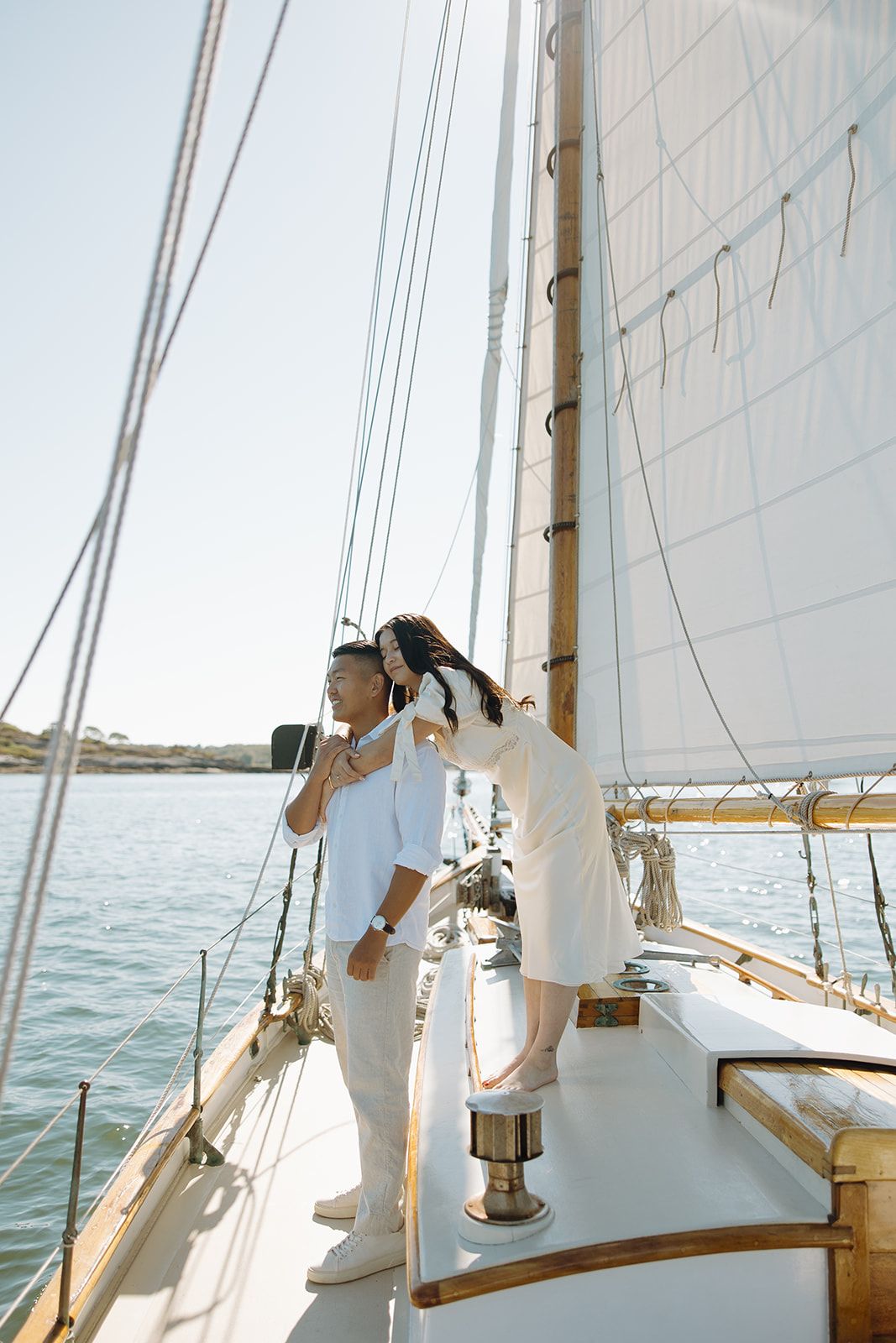 A couple dressed in white outfits stands together on the deck of a sailboat with white sails on calm waters.