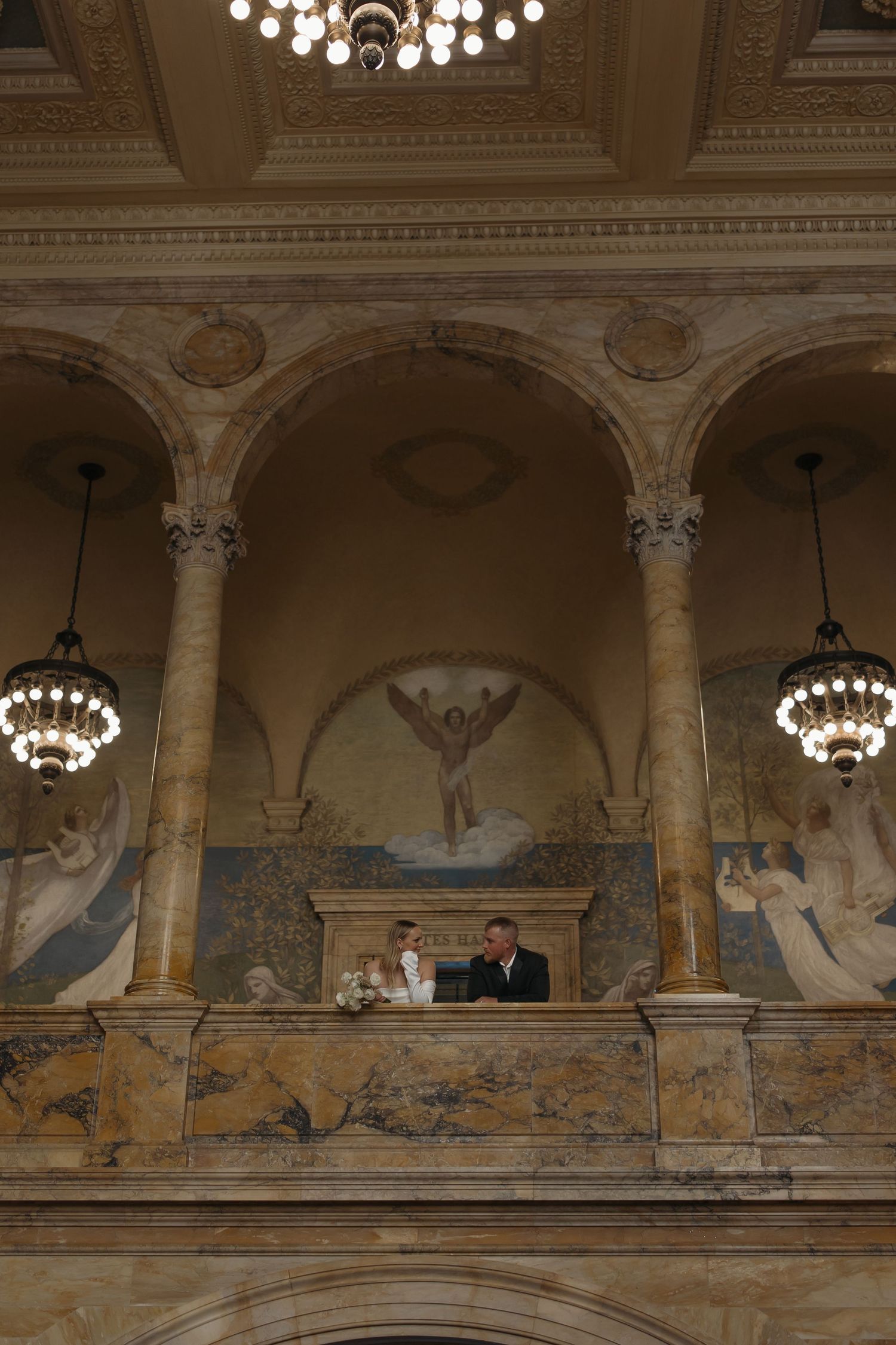 Elegant historic balcony with ornate columns, chandeliers and decorative murals in a grand interior setting.