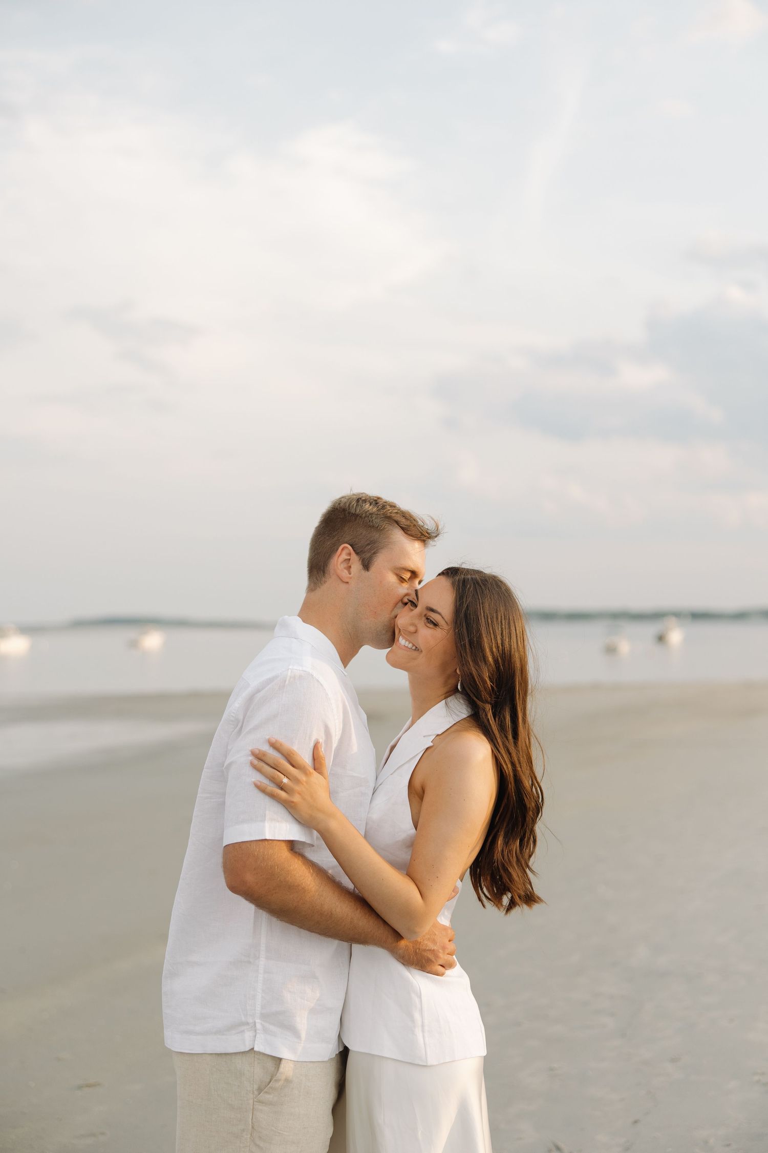 A romantic couple in white outfits sharing intimate kisses on a serene beach at sunset.