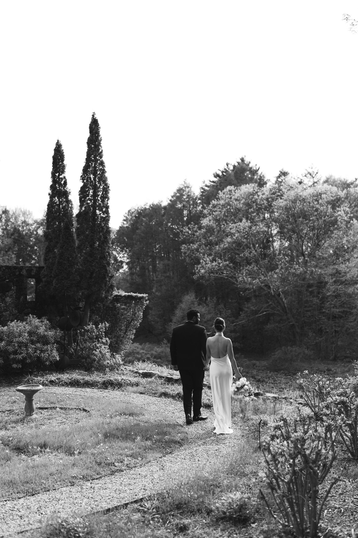 A couple walks along a winding garden path surrounded by cypress trees and natural landscaping in black and white.