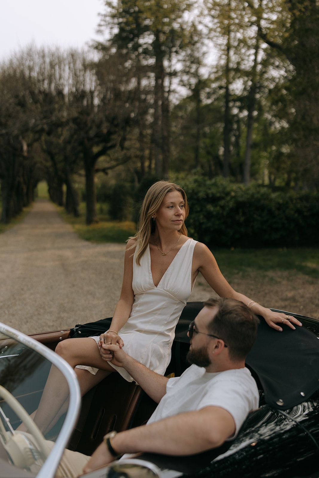 Romantic couple lounging together in a vintage convertible car parked on a tree-lined path during autumn.