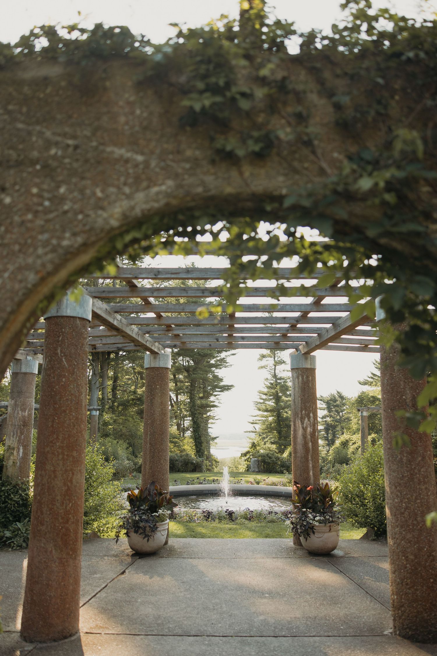 A covered walkway with stone columns and wooden beams creates a shaded path along the waterfront at sunset.