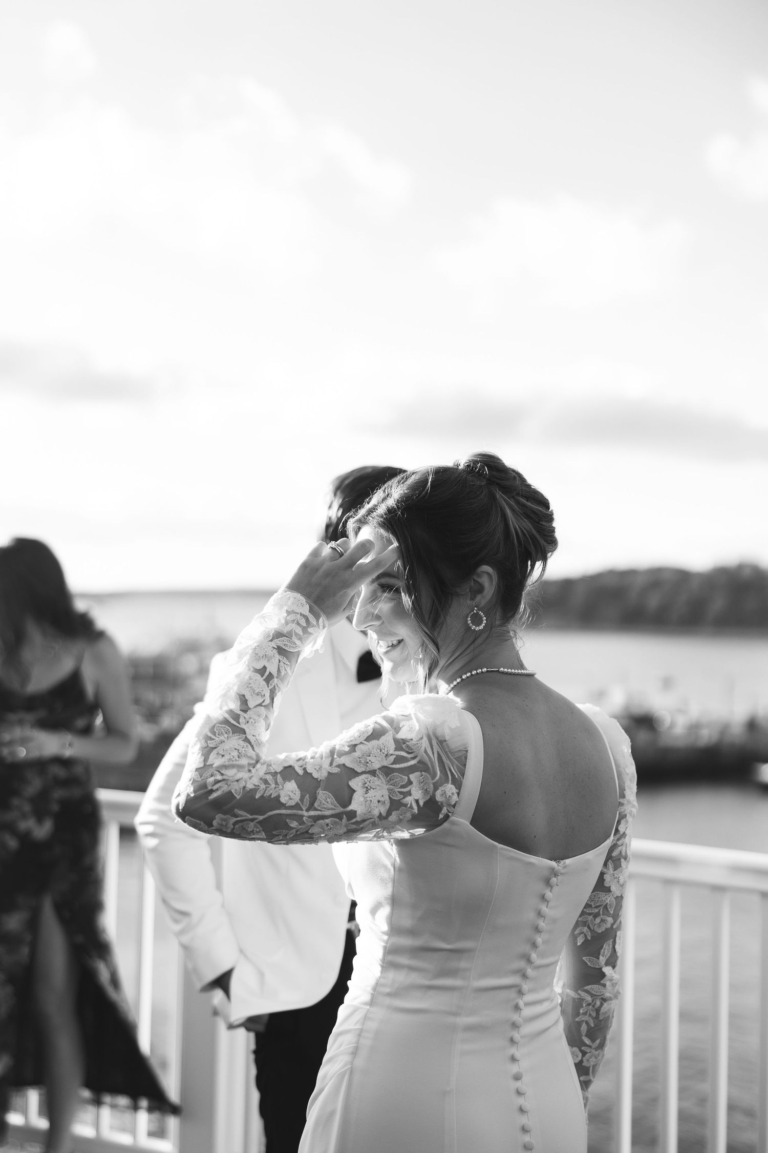 A black and white photo shows a couple in wedding attire standing by a railing overlooking water.