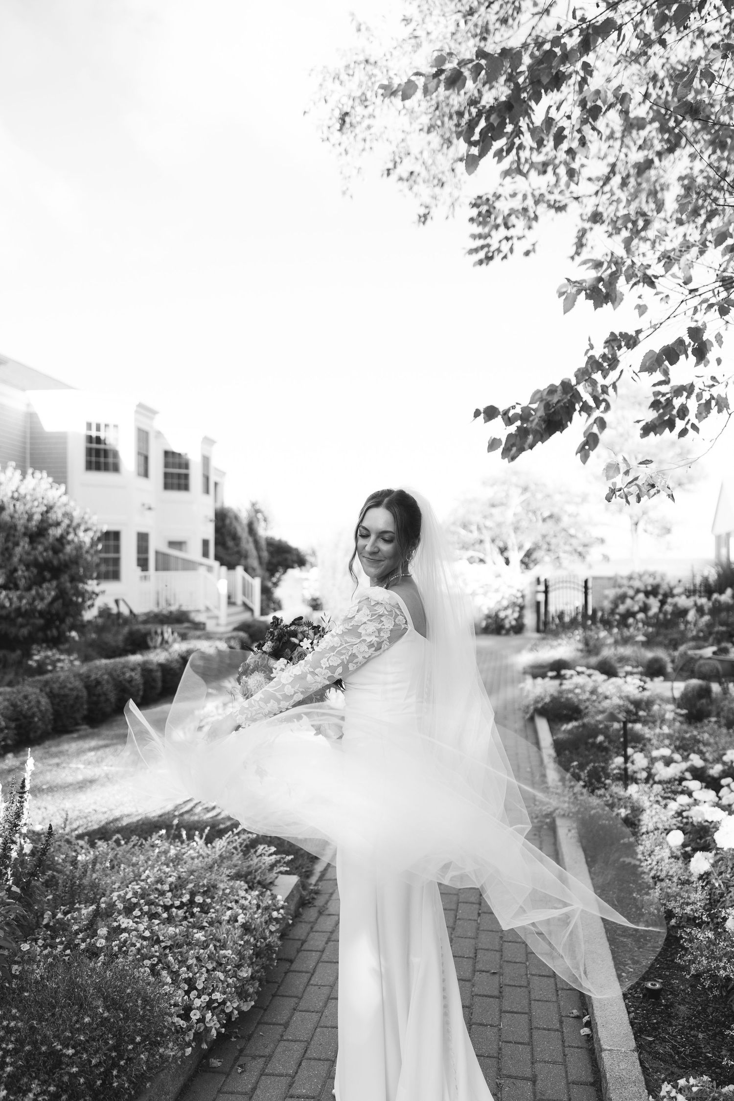 A bride in a flowing white dress twirls gracefully on a brick path surrounded by blooming garden flowers in black and white.