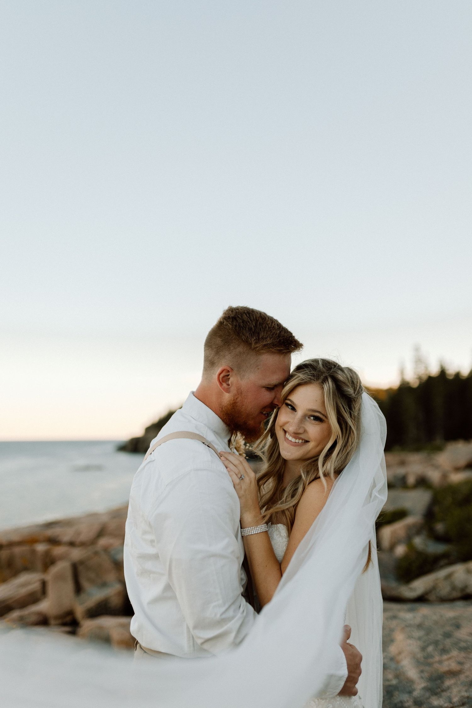 A romantic beach wedding photoshoot at sunset with a couple embracing on rocky coastal shores.