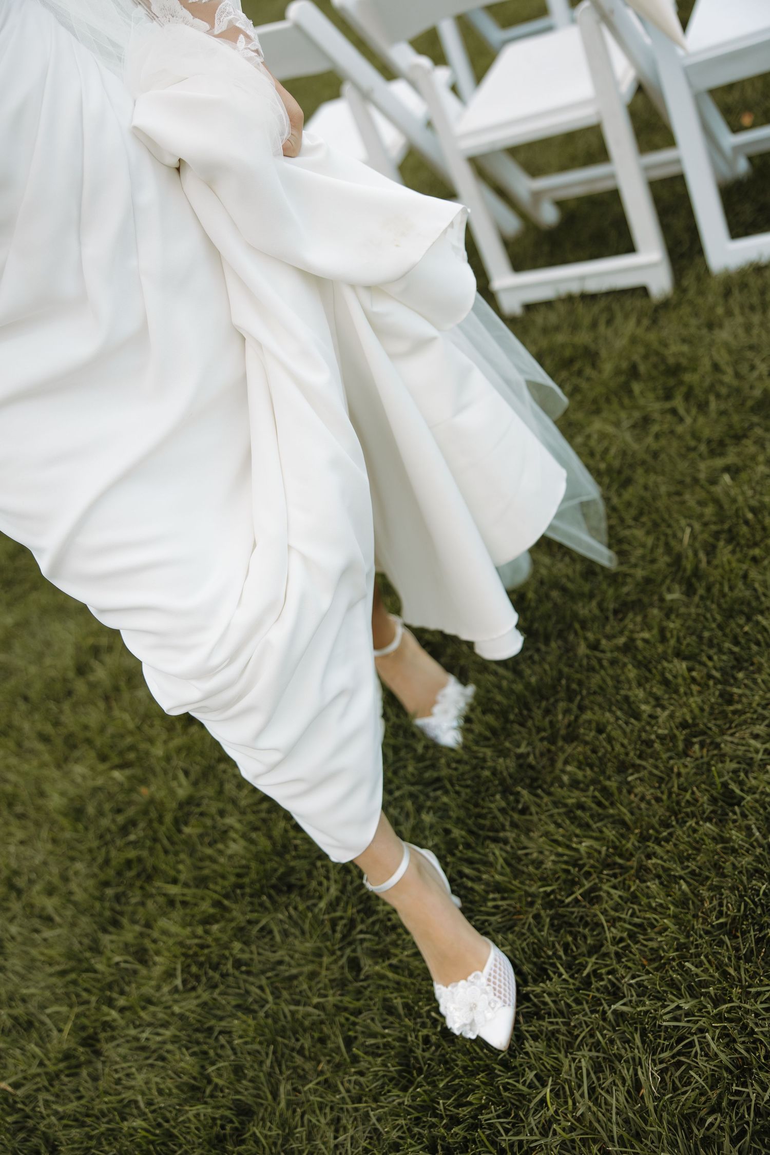 White wedding dress flowing over green grass with elegant white bridal shoes peeking underneath.