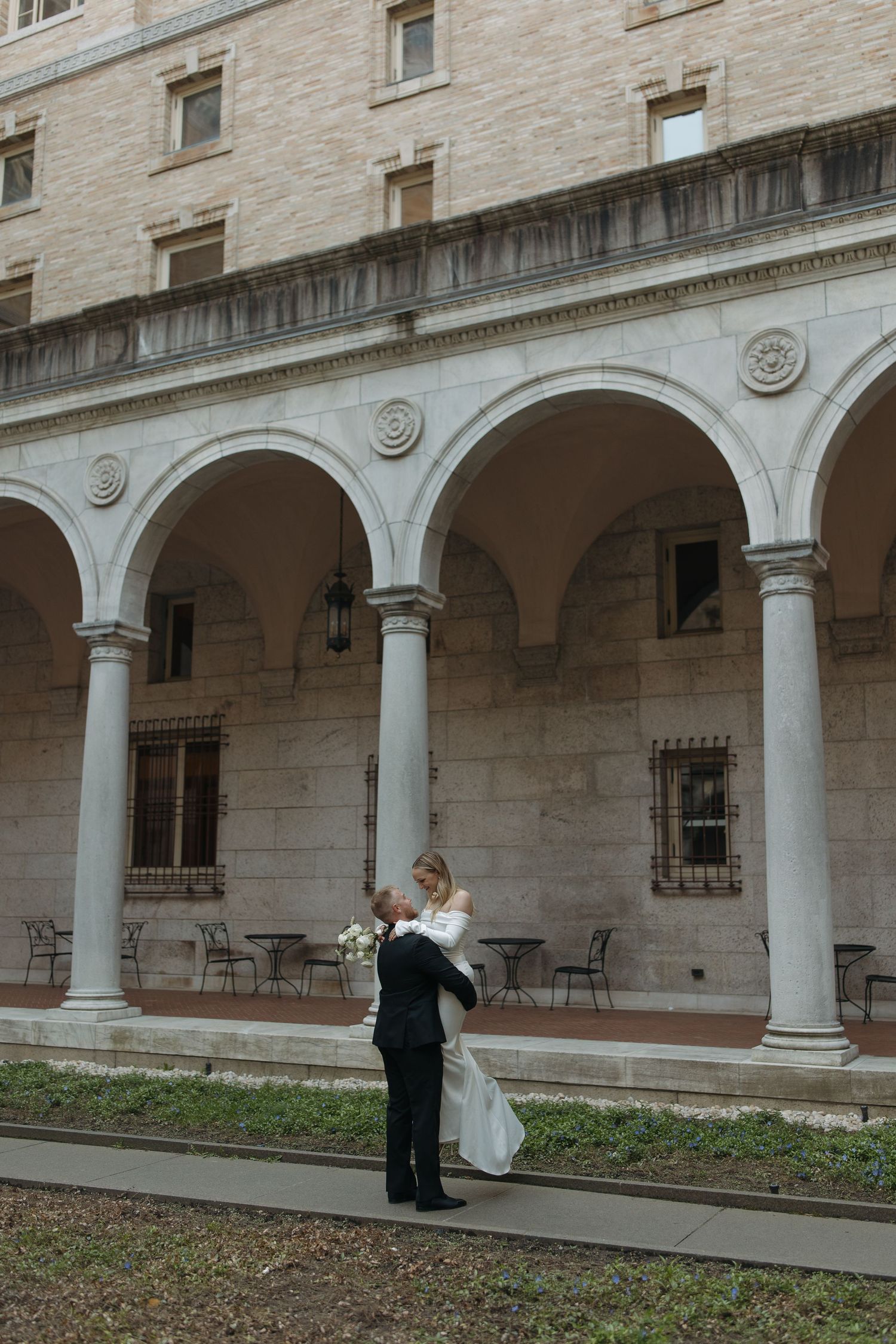 A wedding photo shoot taking place in an elegant stone colonnade with arched pillars and a courtyard garden.