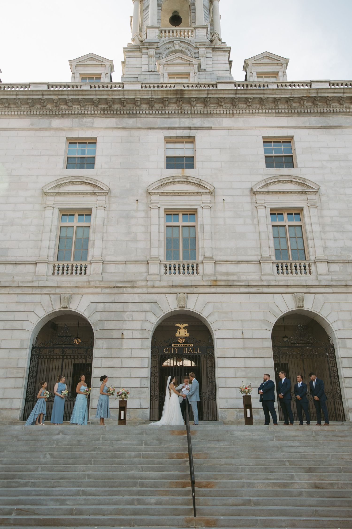 Wedding party stands on grand stone steps of historic courthouse with classical architecture and ornate white facade.