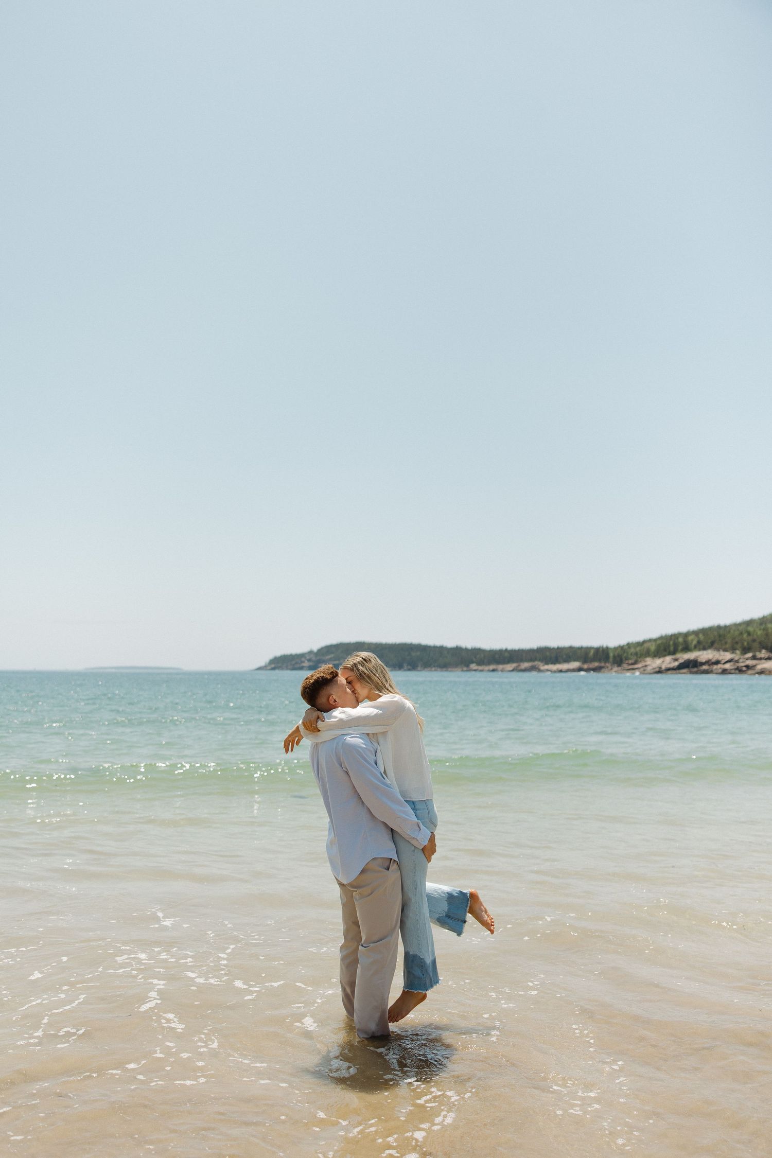 A couple embraces on a sunny beach with turquoise waters and coastal cliffs in the background.