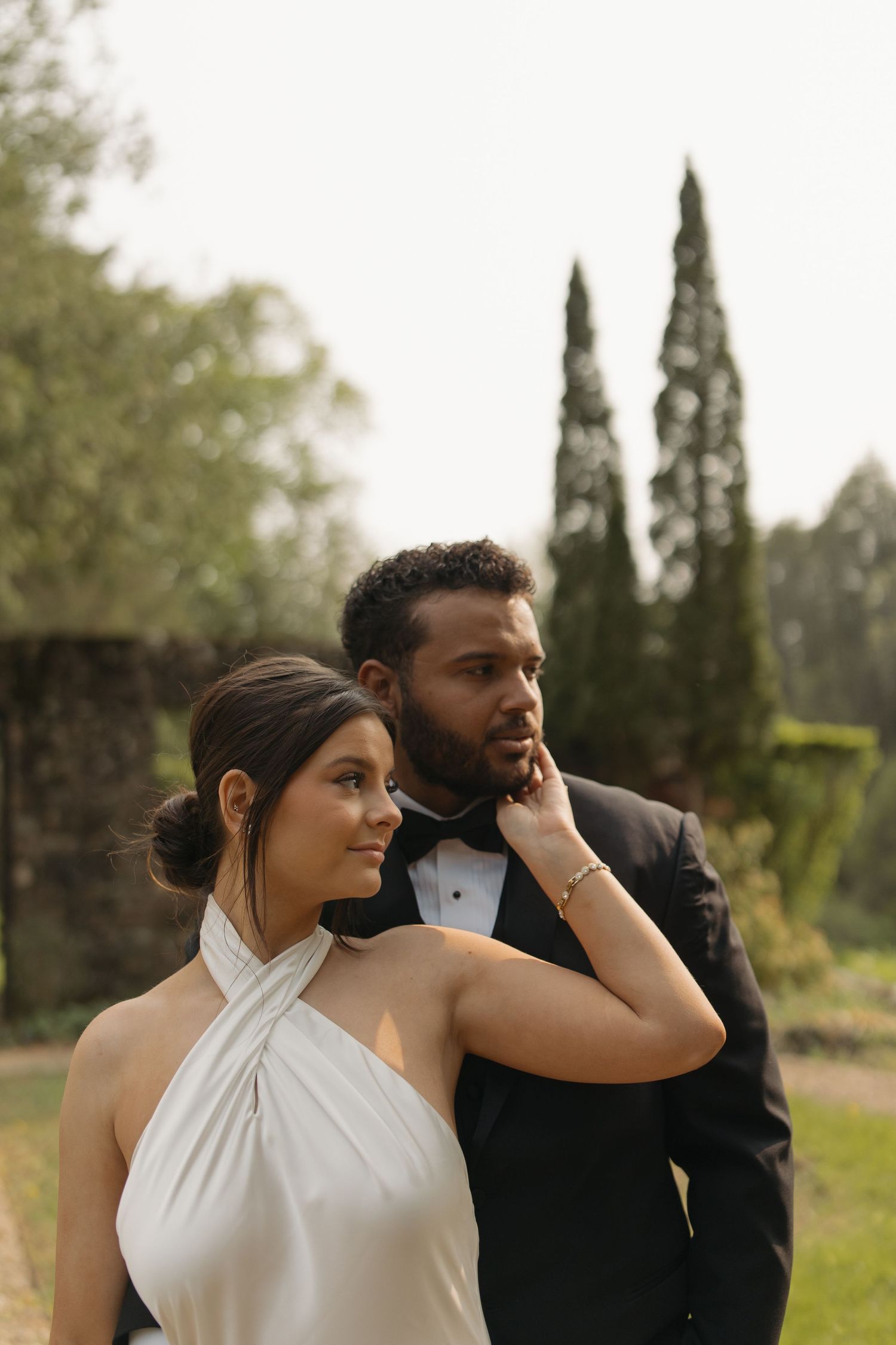 A couple in formal attire poses together in a garden setting with cypress trees in the background.