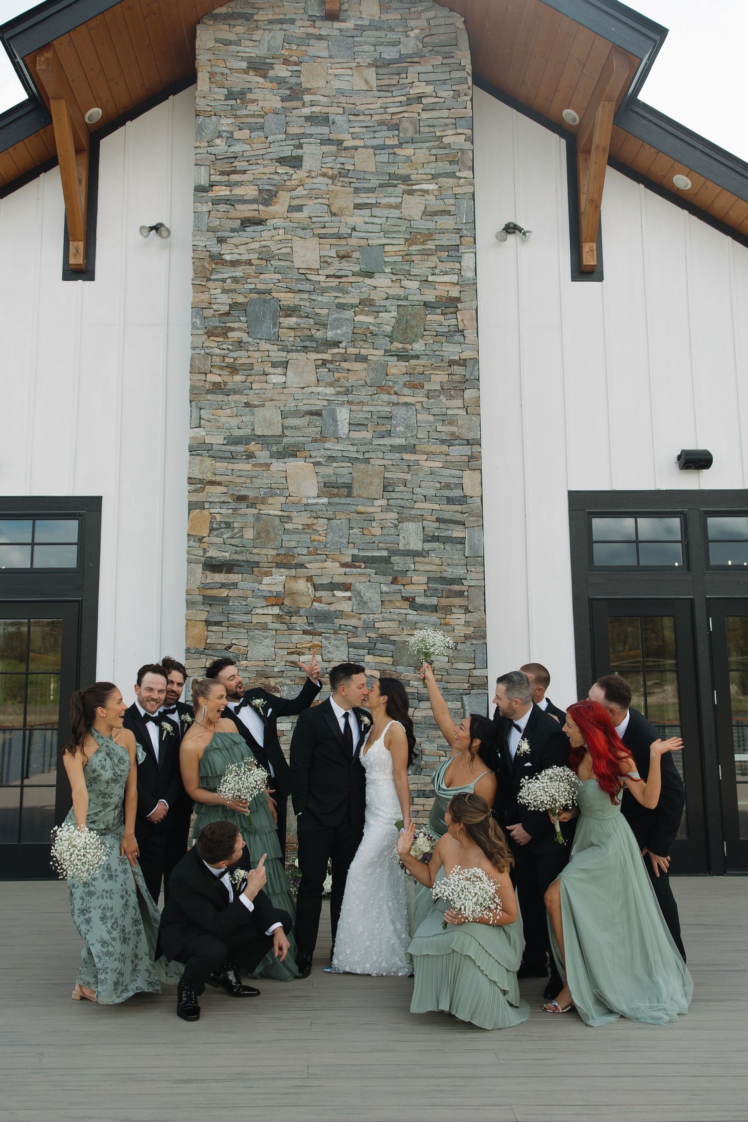 Wedding party in mint green dresses and black tuxedos poses playfully on modern stone and white building porch.