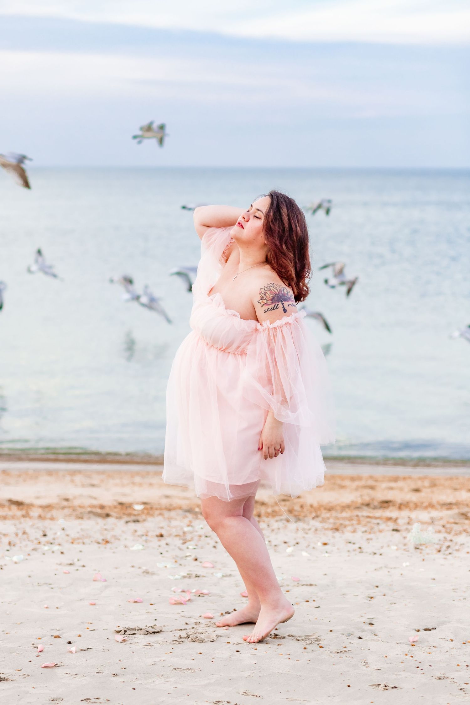A pregnant mom stands on a beach in Norfolk, Virginia wearing a pink dress with birds flying behind her.