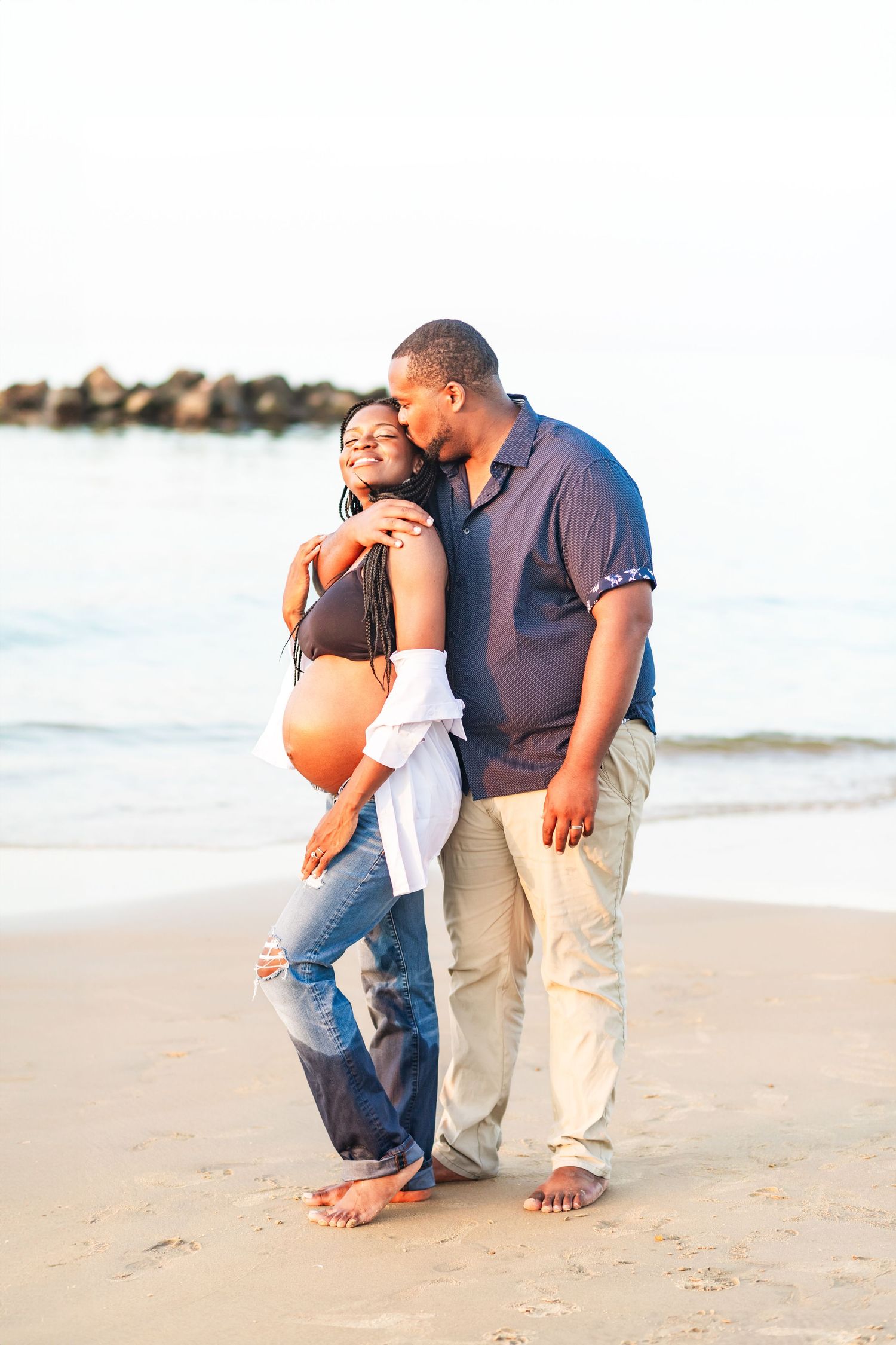 A maternity photo taken at a beach in Norfolk, VA. Dad hugs and kisses mom on the side of her head.