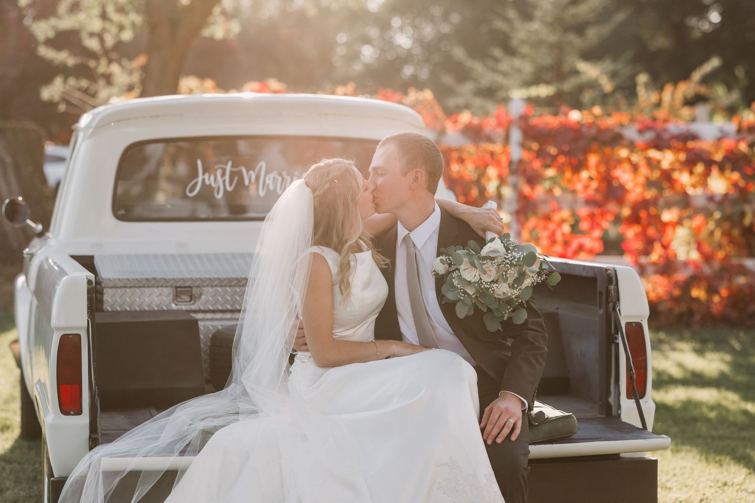 Newlywed couple kissing in the bed of a white vintage truck with a Just Married sign on their wedding day.