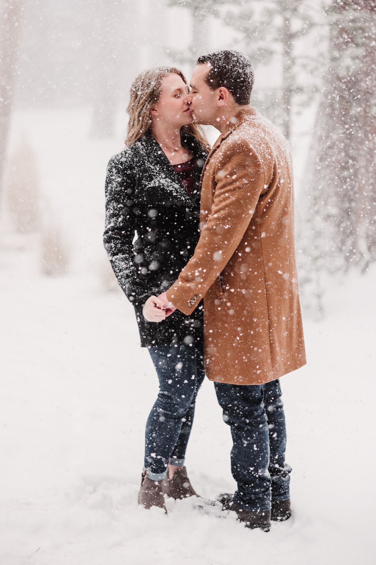 Couple sharing a romantic kiss in the snow, wearing winter coats and jeans during a snowy outdoor engagement photo session.