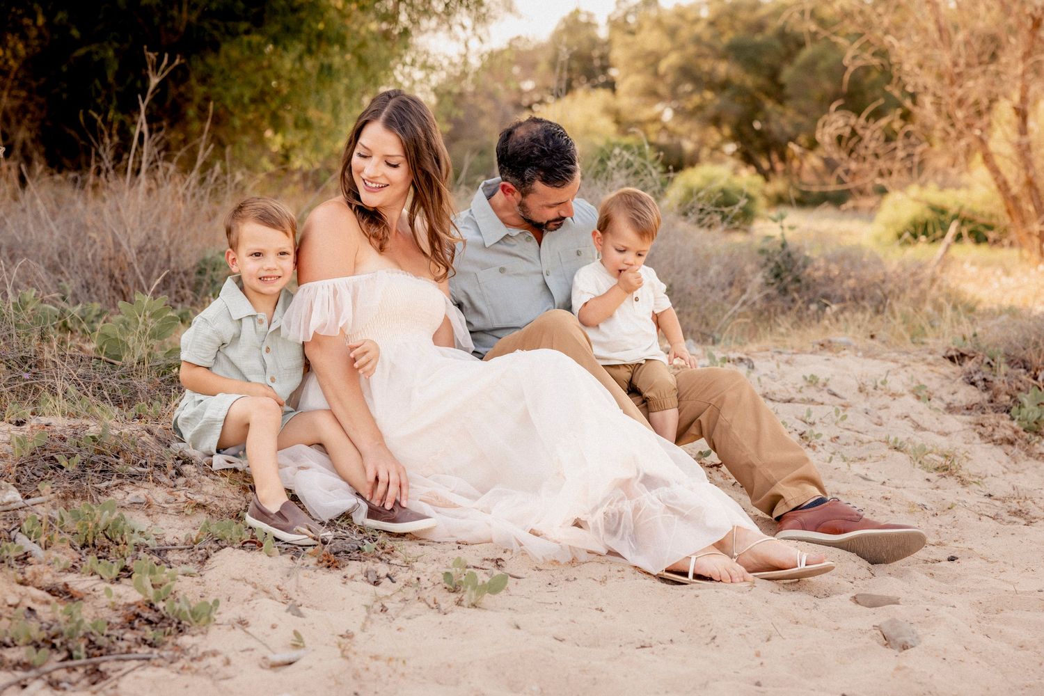 Pregnant woman in white dress sits with husband and two young boys on sandy path surrounded by nature.
