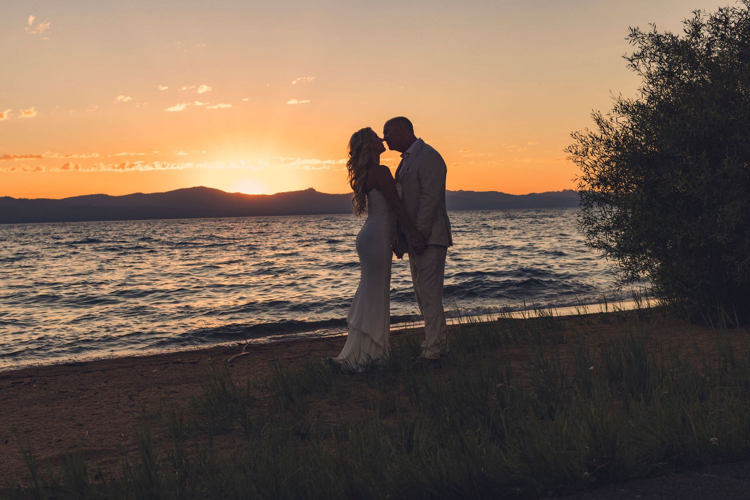 Couple sharing a romantic kiss at sunset by a lake, silhouetted against a golden sky, in wedding attire.