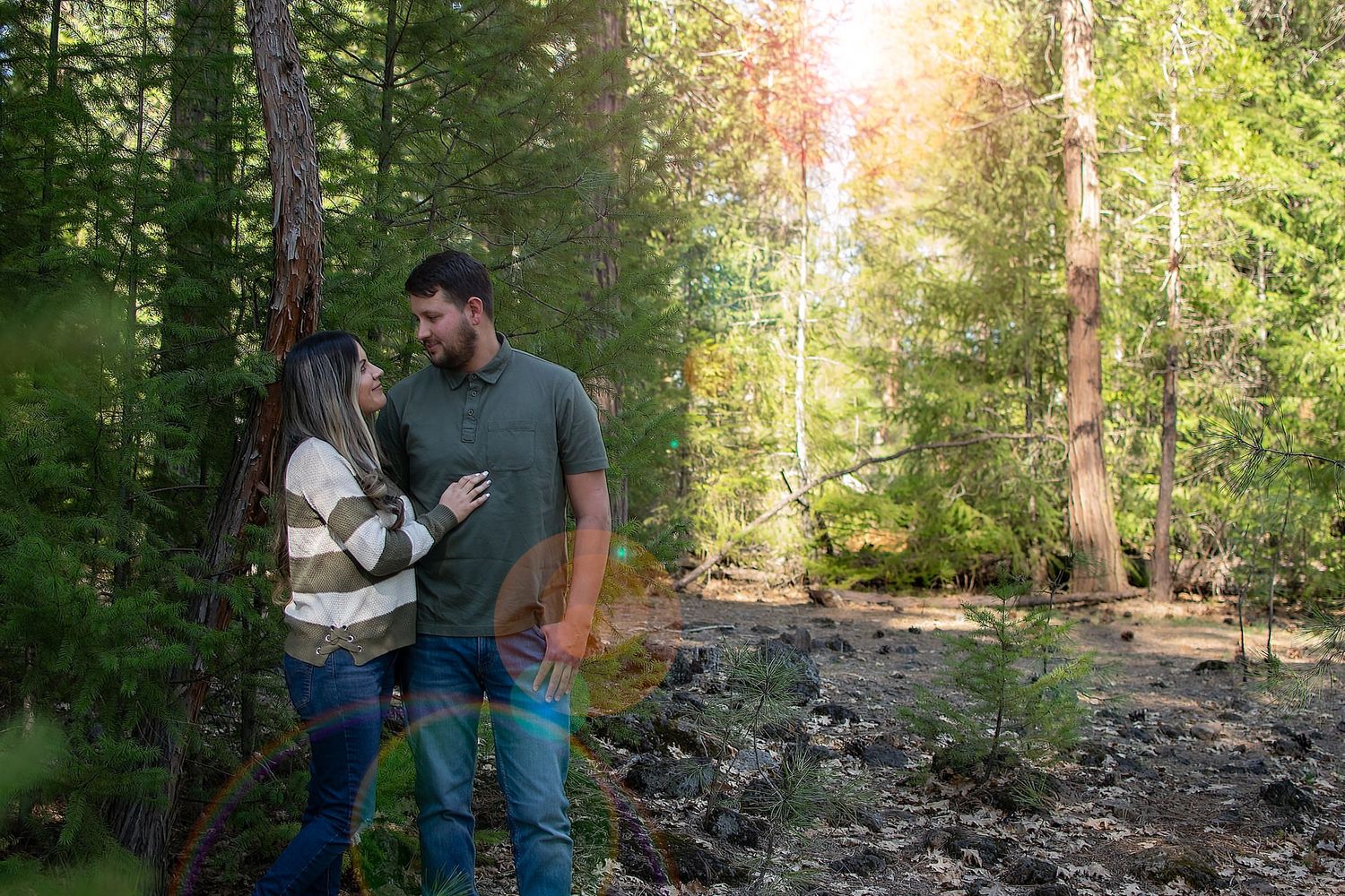 Couple poses in sunlit forest with golden light streaming through trees.