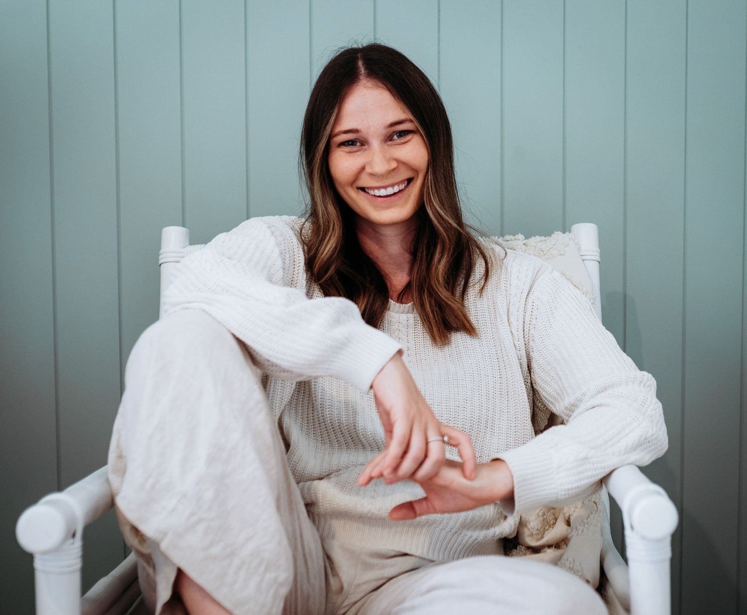 Woman in white sweater sitting on couch with pillow smiling at camera in casual lifestyle photoshoot.