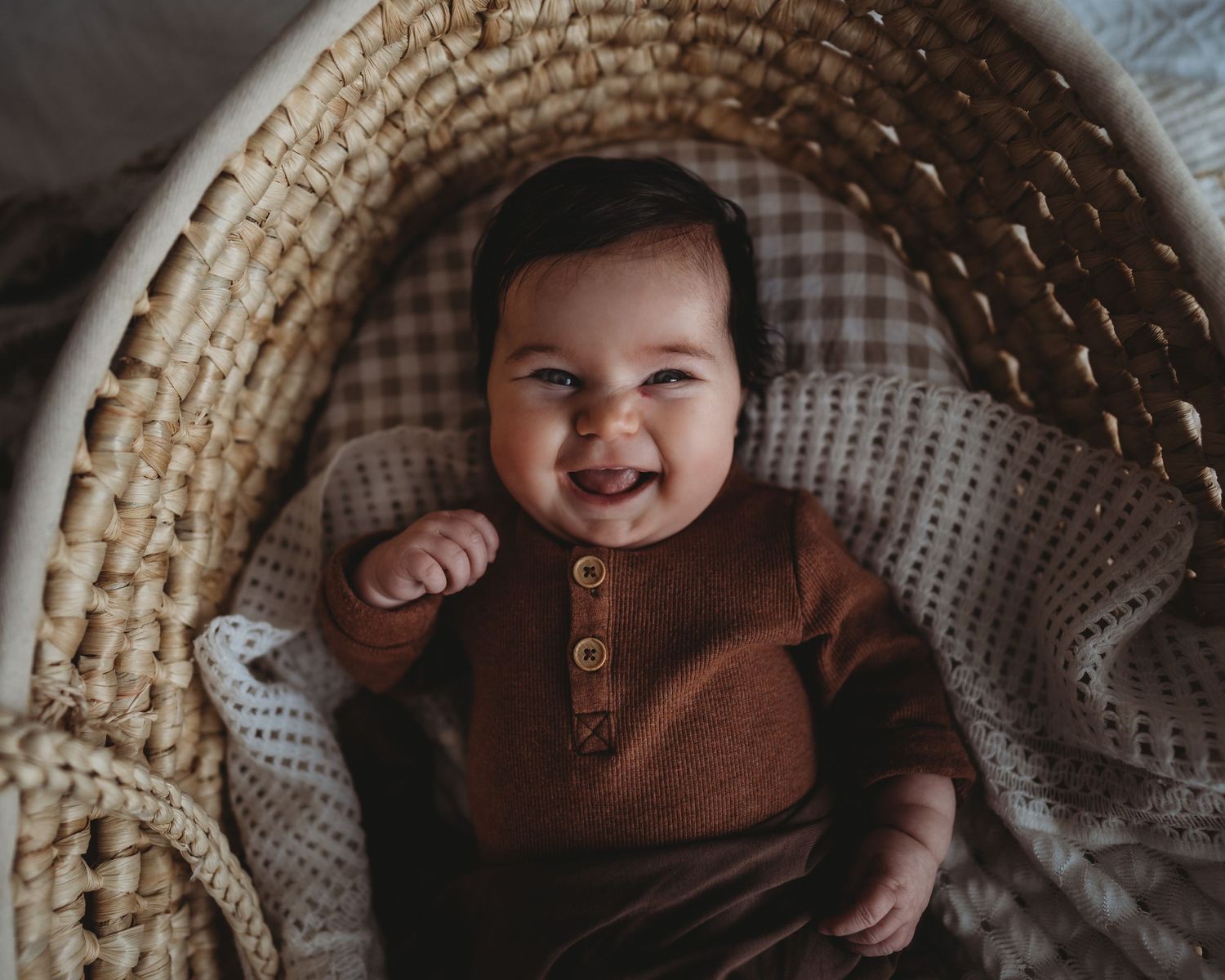 Baby smiling while lying in woven basket wearing brown outfit.