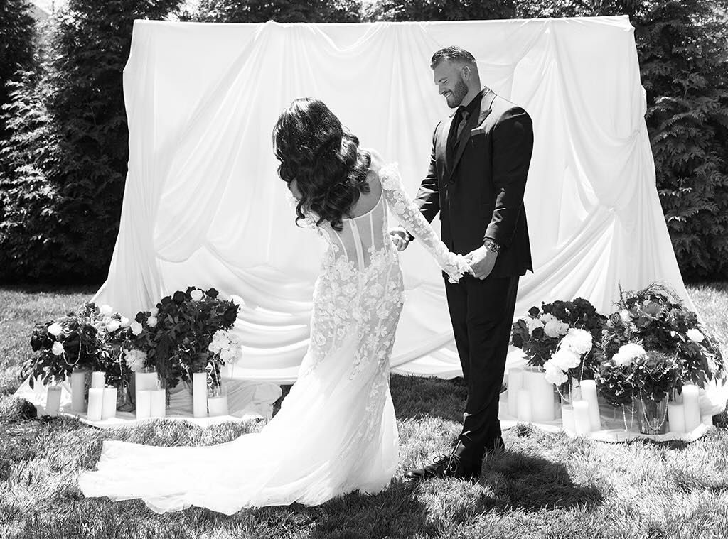 Black and white outdoor wedding ceremony with bride in lace gown and groom holding hands before floral arrangements.