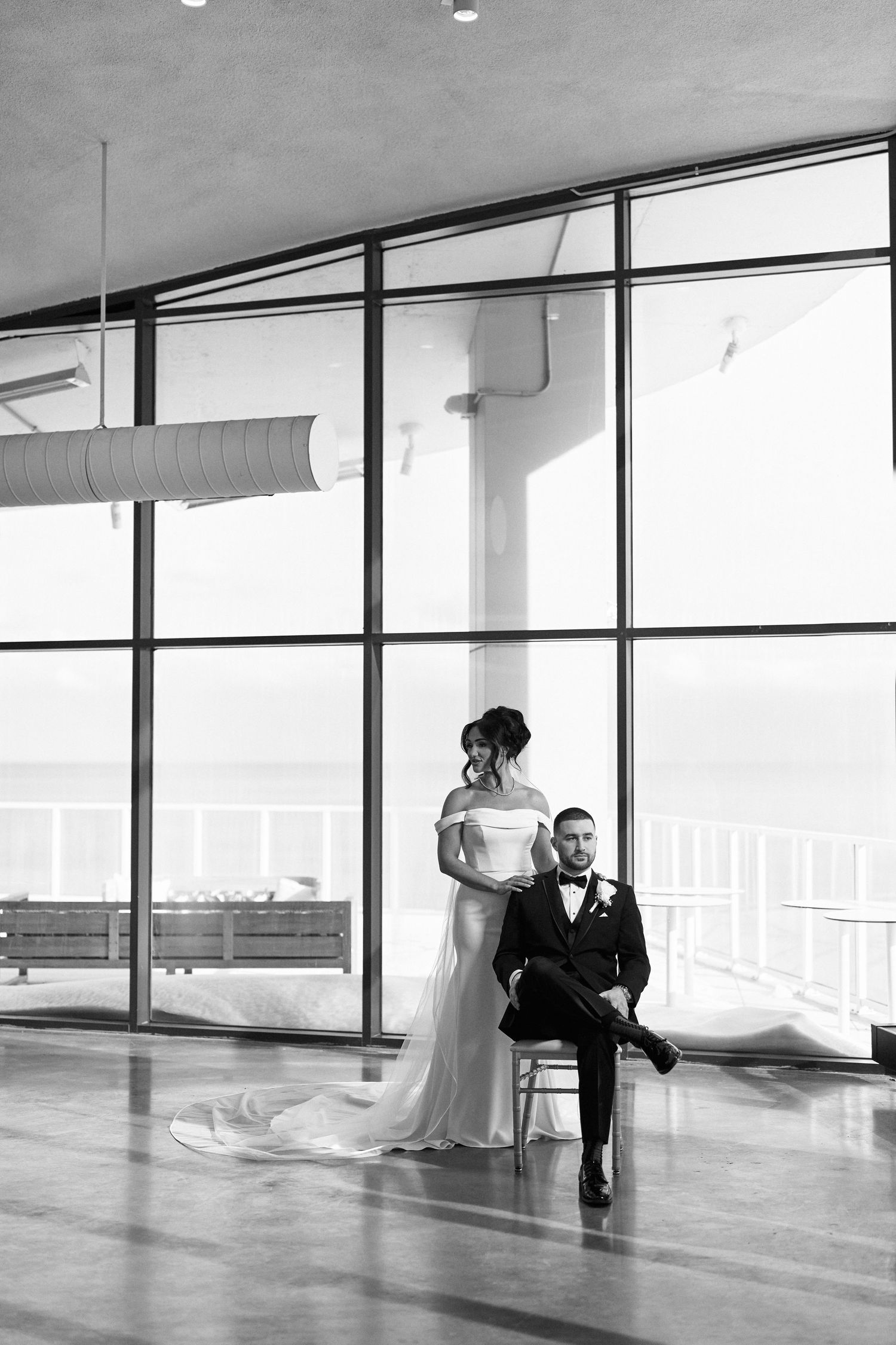 Black and white wedding photo of bride standing behind seated groom before large windows with ocean view.
