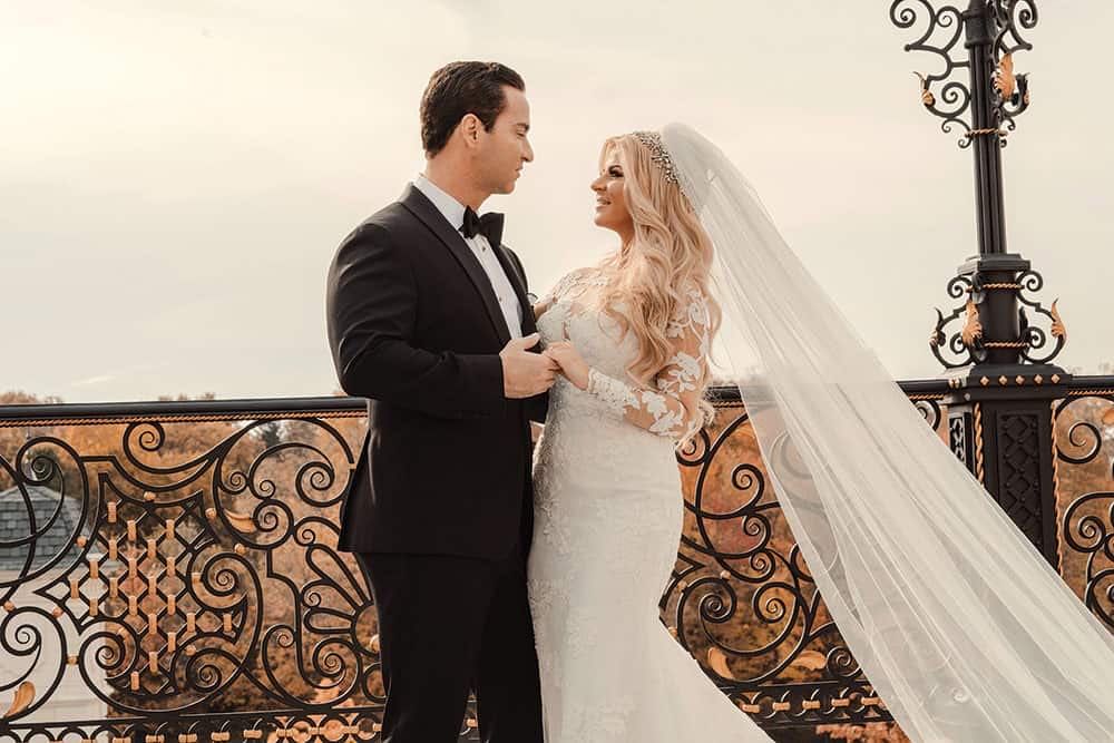 Elegant bride and groom sharing a romantic moment on a balcony in wedding attire.