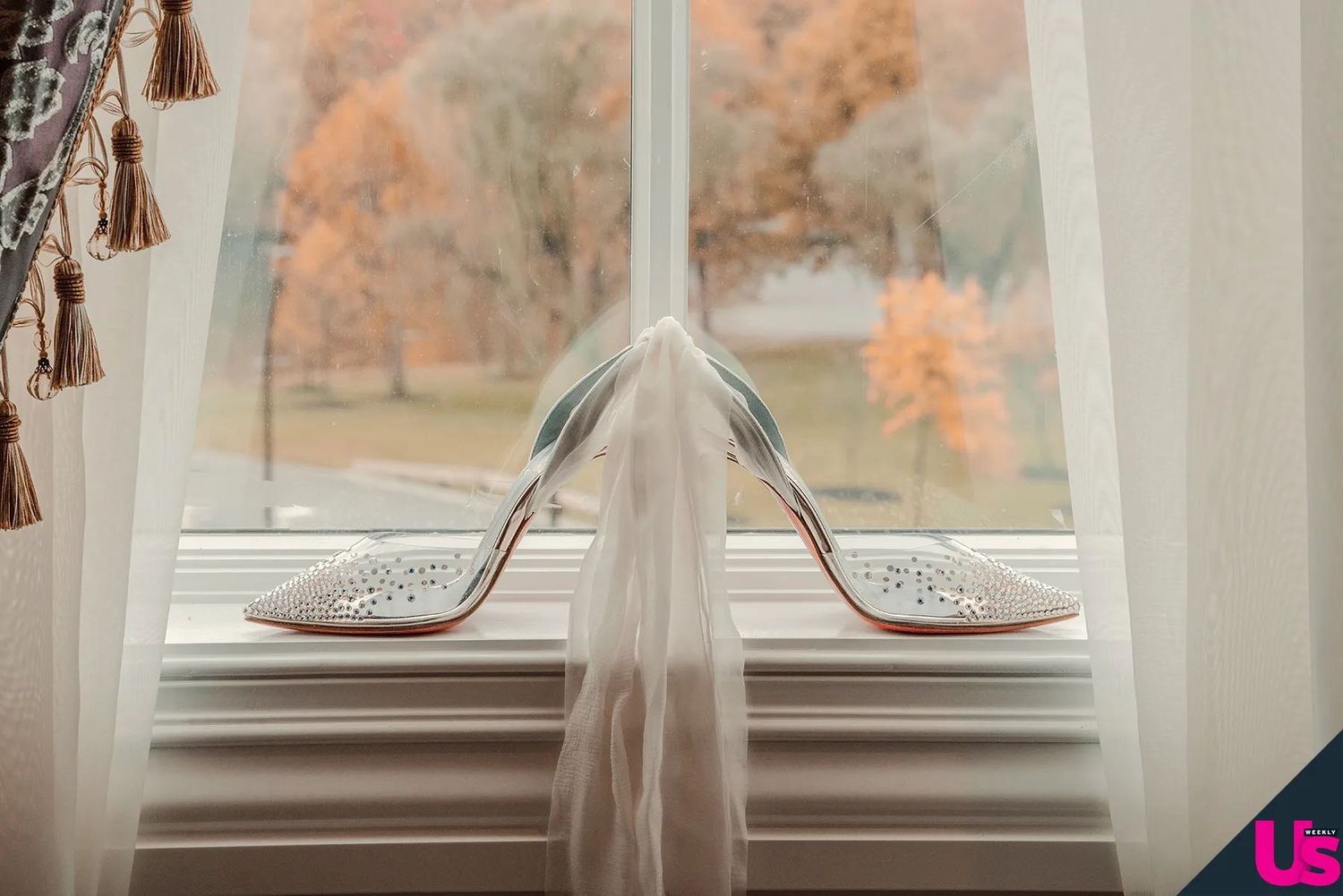 Elegant white Christian Louboutin wedding heels with red soles displayed on a windowsill with a bridal veil and autumn foliage.