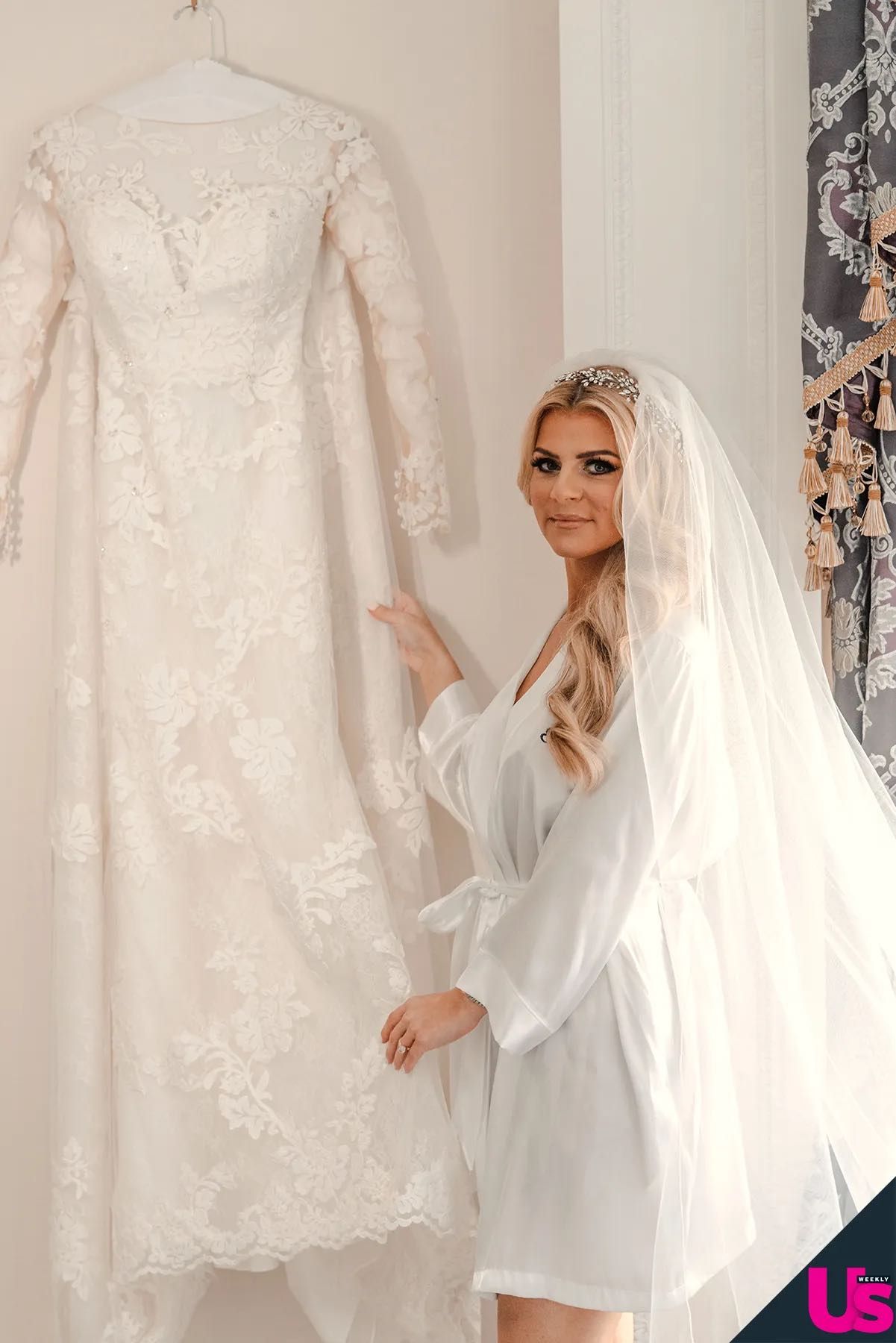 A blonde bride in a white robe and veil admires her lace long-sleeve wedding gown on a hanger indoors.