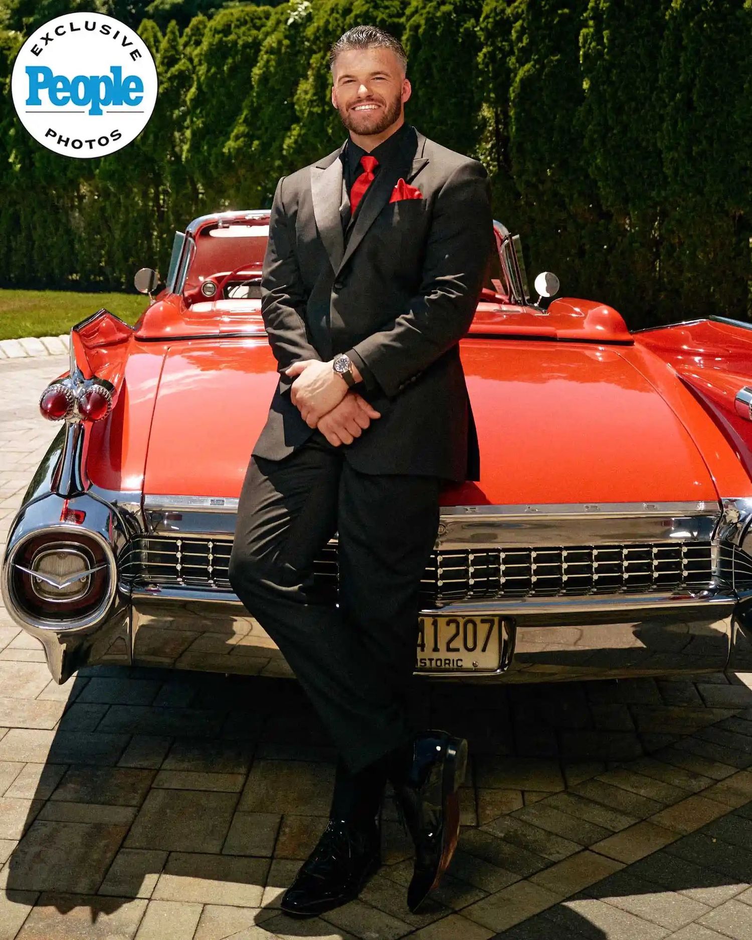 Man in black suit with red tie leaning on classic red Cadillac, featured in People magazine exclusive photos.